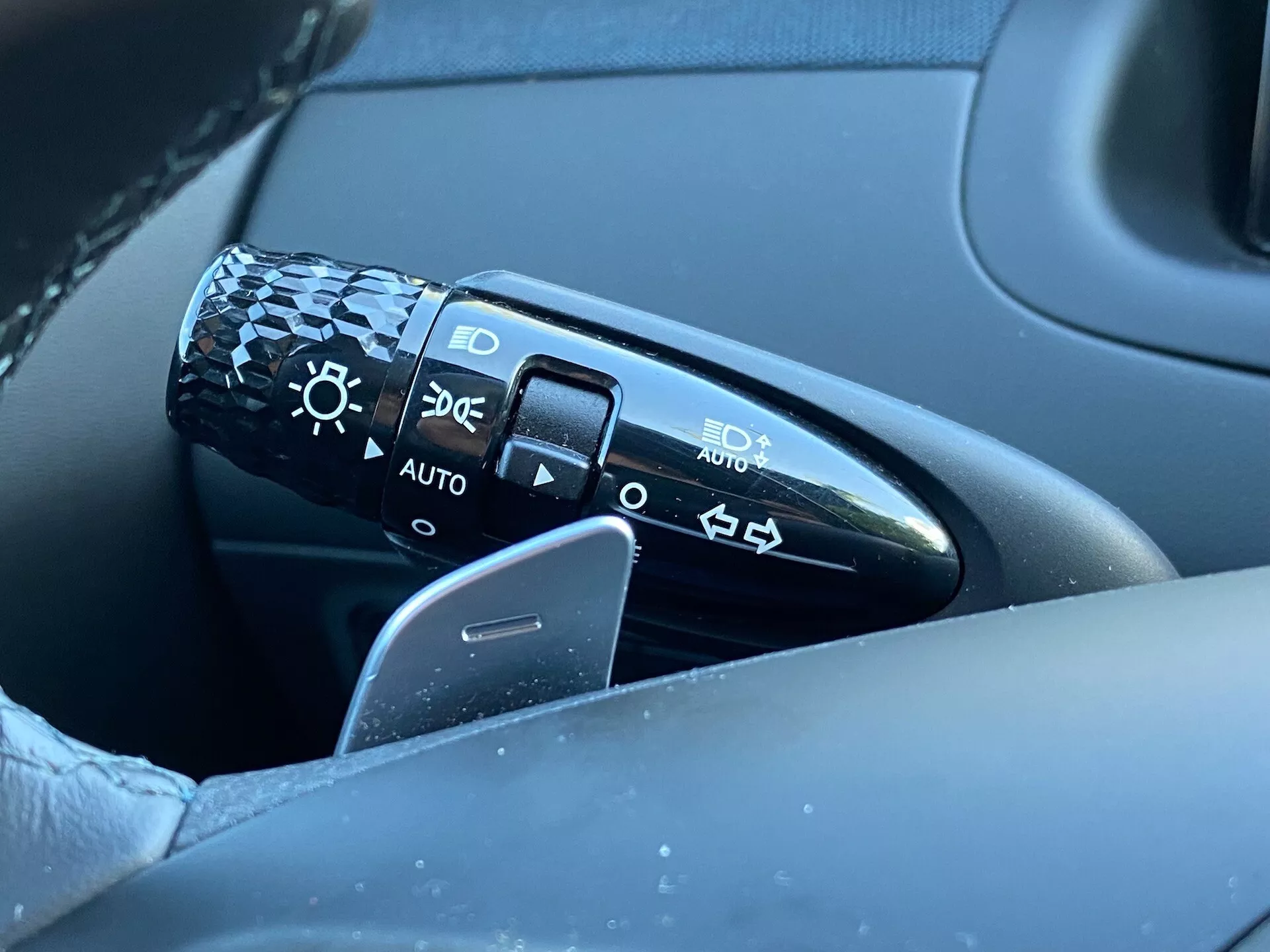 Közelkép a Hyundai Tucson 2020 kormányoszlopán a kormánykeréktől balra található vezérlőkaról. A karon látható szimbólumok a fényszórók beállításait jelzik: kikapcsolva, automatikus fényszórók, parkolófények és alacsony/magas fényszórók. A kép a kart, a kormánykerék egy részét és a vezetőoldali ajtó részét egy elmosódott háttérrel mutatja, kiemelve a részleteket és a minőségi designt. Az 'AUTO' pozíció automatikus fényszórórendszert jelez.