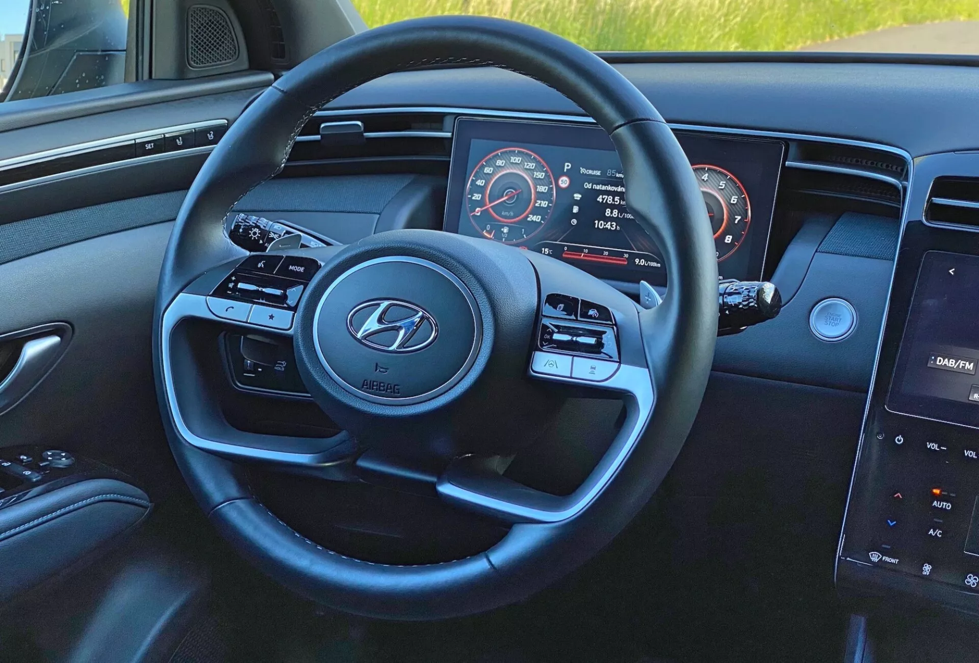 A Hyundai Tucson 1.6 T-GDI 2020 belső nézete a sofőr szemszögéből, a modern háromküllős kormánykerékre fókuszálva, Hyundai logóval, média- és sebességtartó automatika vezérlőgombokkal, részben látható műszercsoporttal, amely tartalmazza a sebességmérőt és a fordulatszámmérőt, és a műszerfalon lévő központi érintőképernyős infotainment rendszerrel.