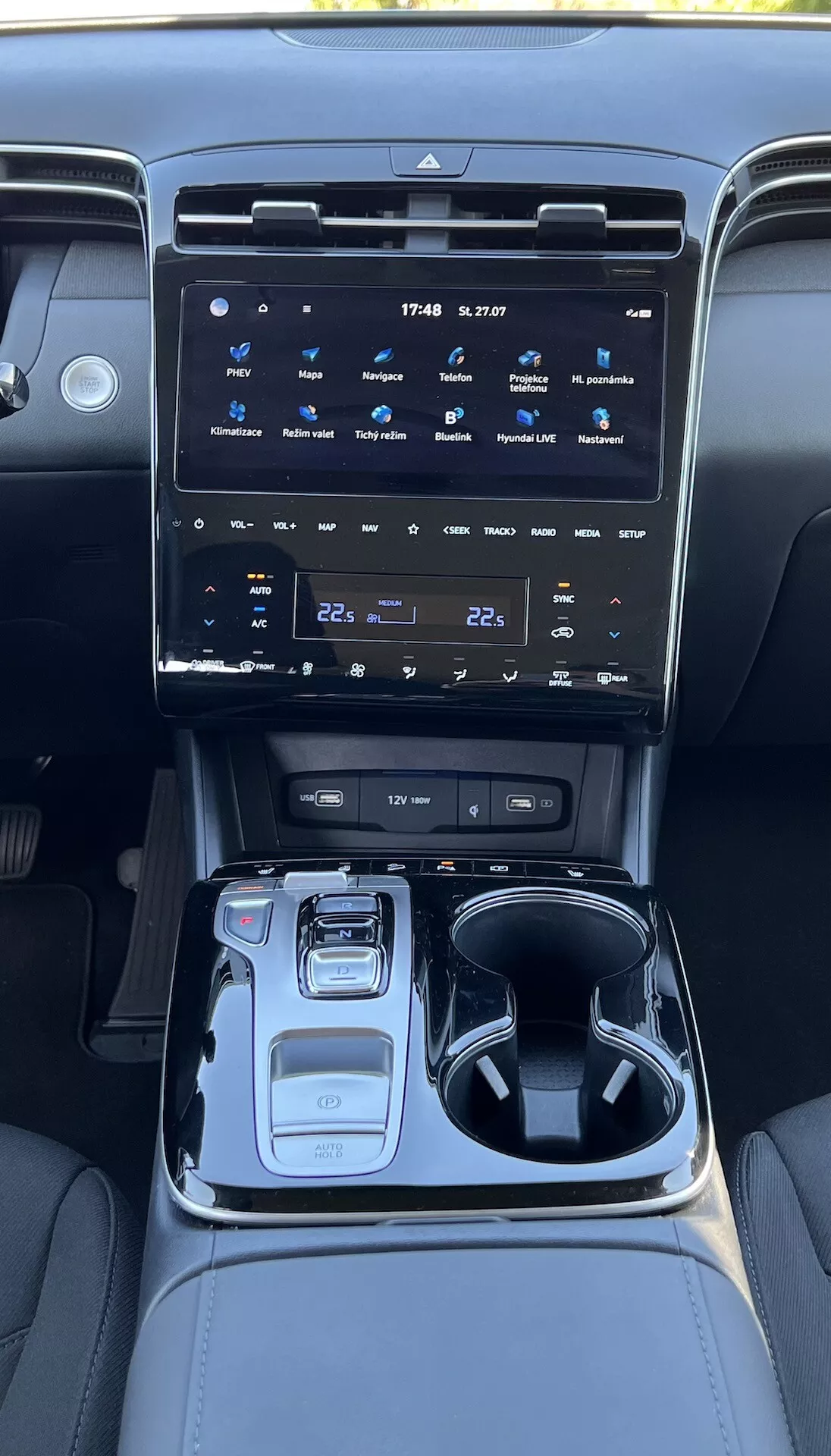 2020-as Hyundai Tucson belső terét bemutató középkonzol egy nagy érintőképernyős infotainment rendszerrel, klímakontroll gombokkal, elektronikus váltógombbal, vezetési mód választóval, pohártartókkal, tároló rekesszel és hangulatvilágítással.