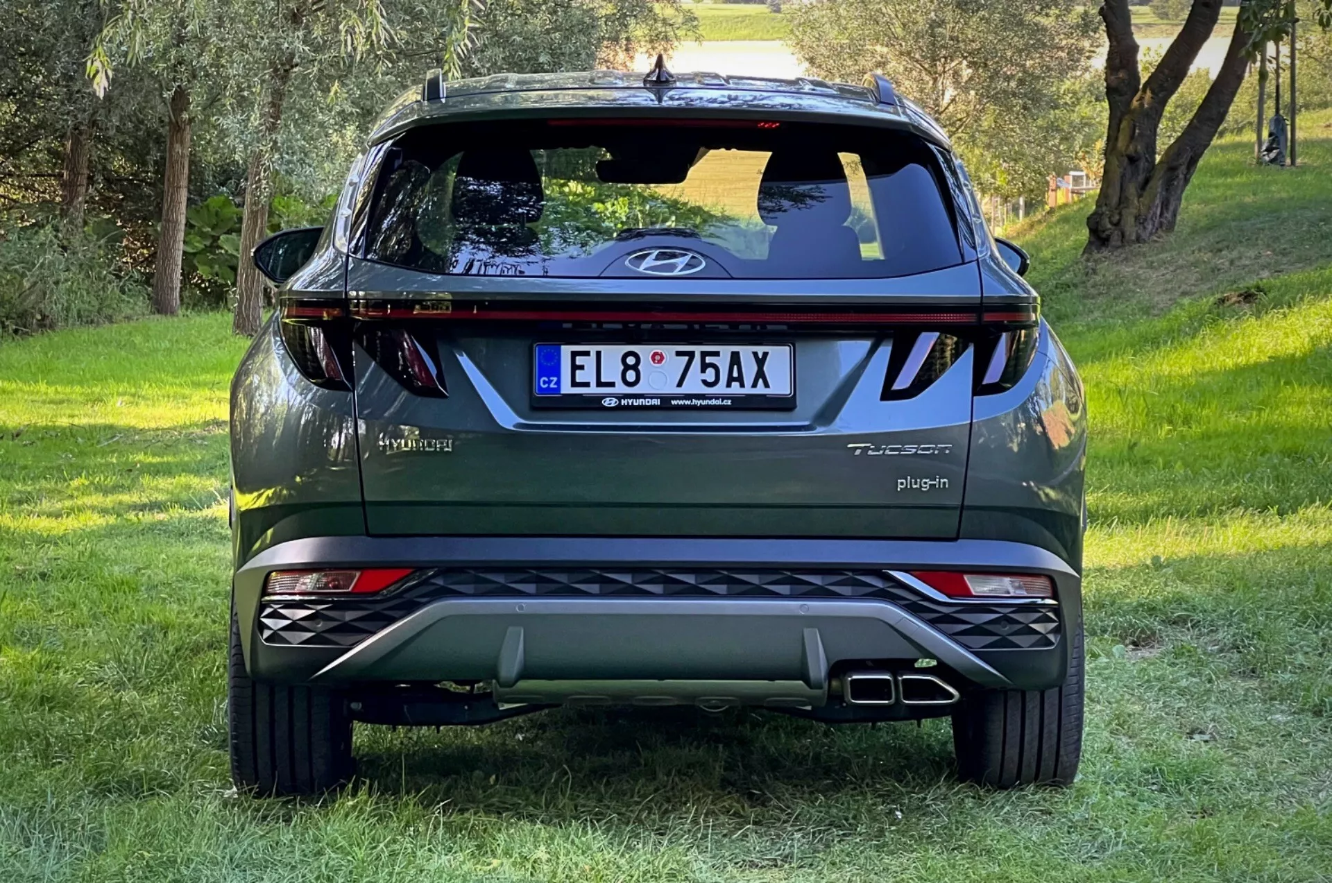 2020-as Hyundai Tucson SUV hátsó nézete füves lejtőn erdős háttérrel, bemutatva a csomagtartó logóját, Tucson és Hybrid jelvényeket, Plug-in feliratot, visszaverő fényeket, dinamikus hátsó lámpákat, hátsó ablaktörlőt, magasan elhelyezett féklámpát, kettős kipufogócsöveket, ezüst futóműlemezeket és AWD jelvényt.