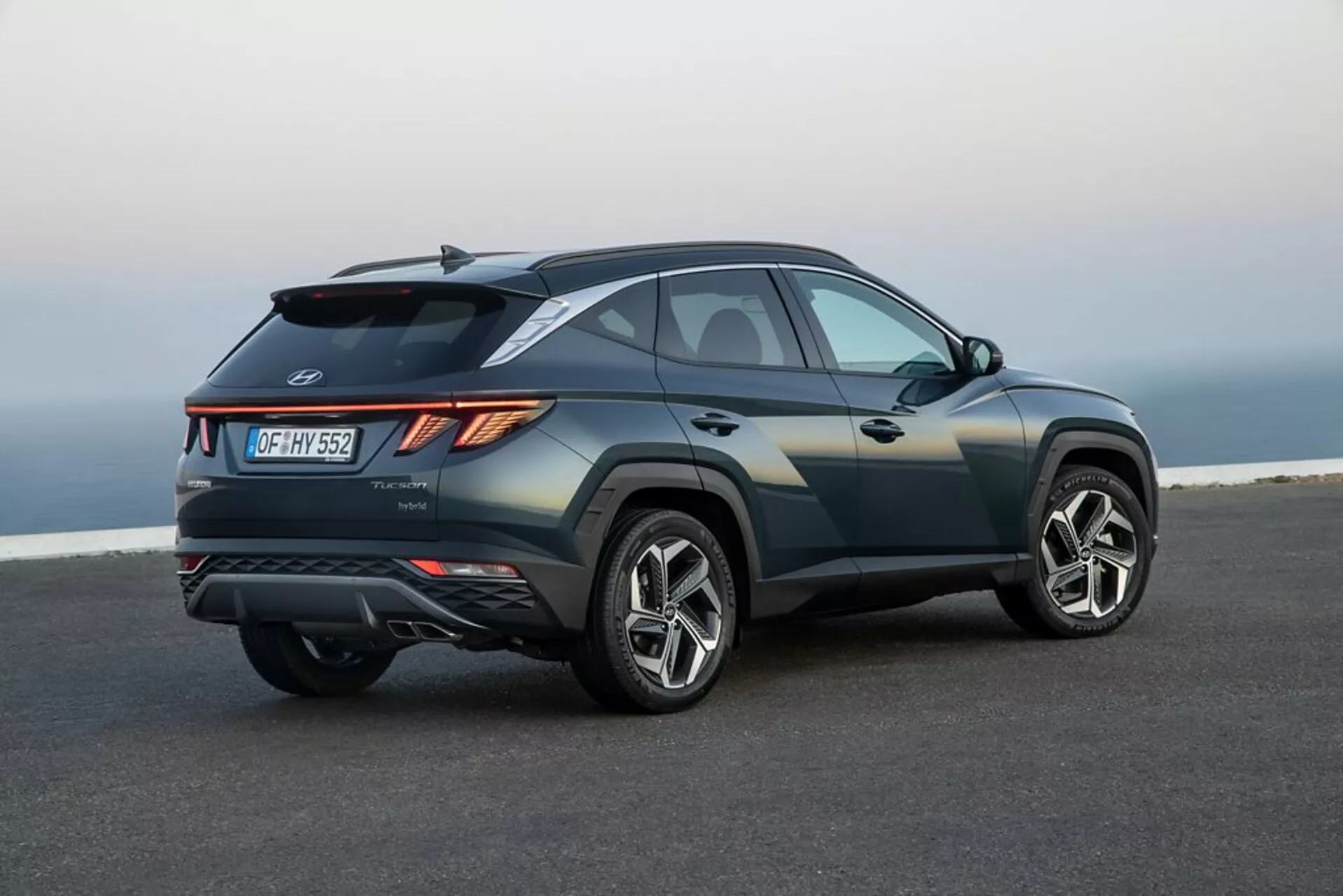 2020-as Hyundai Tucson SUV parkol sima aszfalton, tengerparti vagy emelkedő táj hátterével, bemutatva az elegáns, sportos kialakítását, emelkedő övvonalát, lejtős tetővonala, éles karosszériahajlításait, szögletes LED hátsó lámpáit, hátsó légterelőjét, integrált diffúzorát, kettős kipufogóját és nagy ötvözött kerekeit