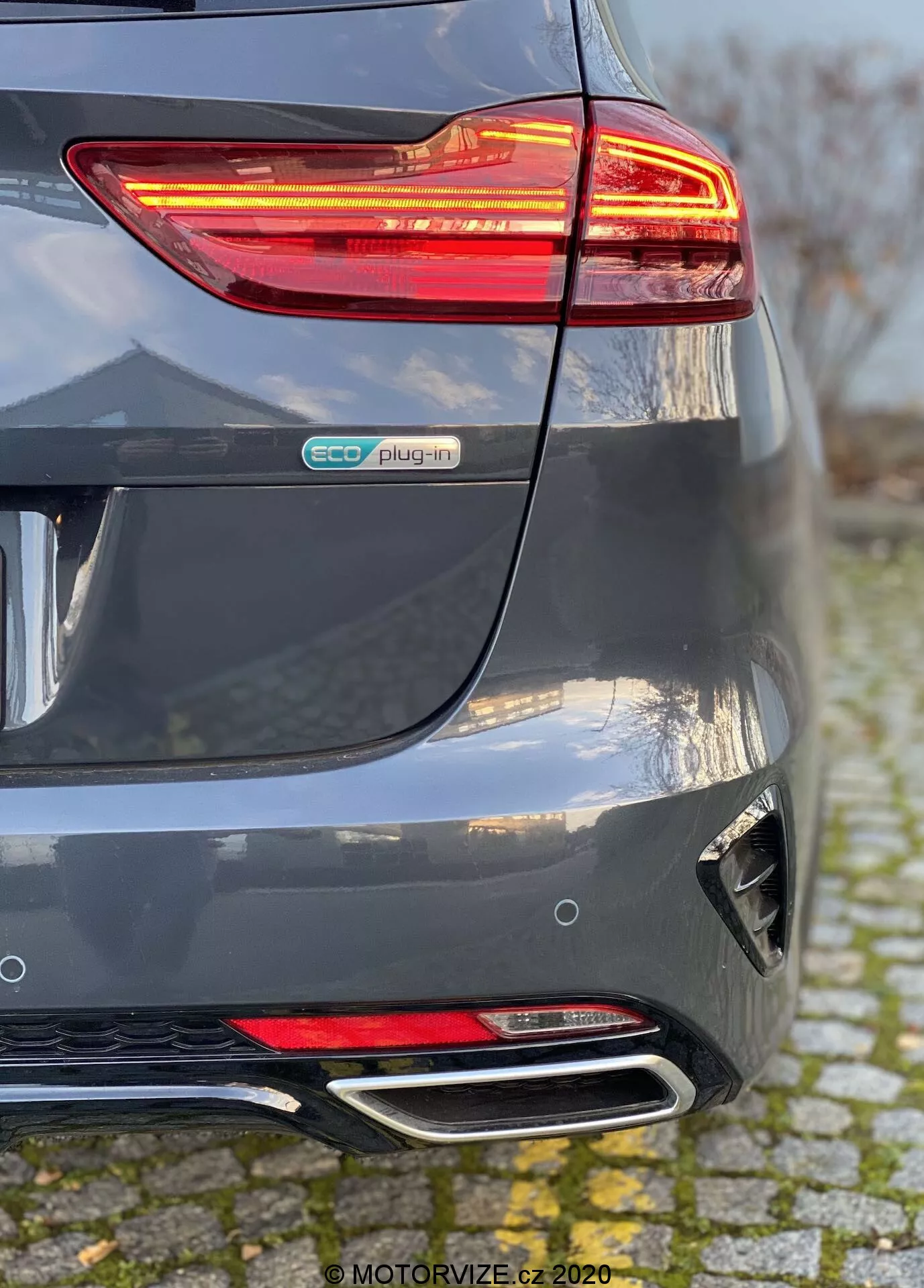 Részletes nézet egy 2018-as Kia Ceed hátsó részéről, amely fémes szürke színű, kiemelve a megvilágított LED hátsó lámpát, az eco plug-in jelvényt, a reflektorral és diffúzorral ellátott kialakított hátsó lökhárítót, valamint a kettős krómezett kipufogóvégeket. A perspektíva a hátsó lökhárító és a jobb oldal találkozását mutatja, hangsúlyozva a hibrid elektromos jármű elemeit és a modern dizájn jellemzőit.