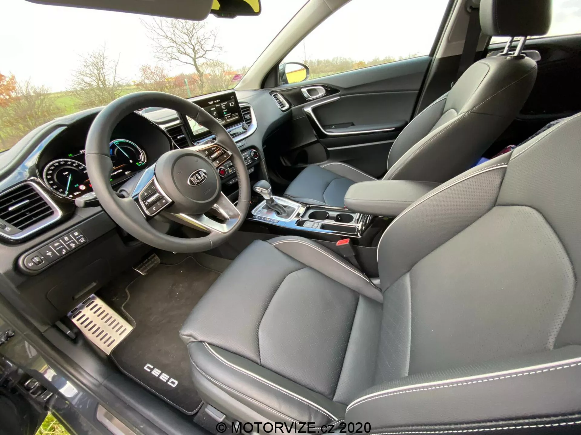 A Kia Ceed 2018 belső fotója az utas szemszögéből, egy szögben fényképezve, amely hangsúlyt fektet a műszerfalra, a középkonzolra és a vezetőülésre. A kormánykerék több vezérlőgombbal és a Kia logóval jól látható, valamint részben látható a digitális műszerfal is. A műszerfal közepén egy nagy érintőképernyős szórakoztató és információs rendszer található, melyet manuális klímavezérlők vesznek körül. A középkonzol tartalmazza a sebességváltókart, pohártartókat és vezérlőgombokat. Az elülső utasülés bőr kárpitozást kapott, a keverék anyagú ajtópanelek pedig luxus szintet sugallnak. A fém fék- és gyorsító pedálok prémium sportos érzetet kölcsönöznek.