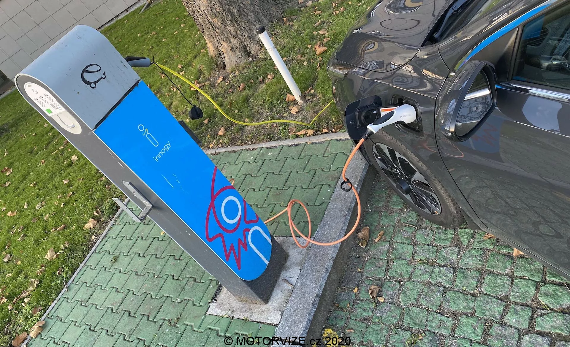 Magas szögből készült kép egy modern elektromos járműről, amelyet egy nyilvános töltőállomáson töltenek. A kép a jármű jobb hátsó részét mutatja, a töltőnyílásra illetve a vastag narancssárga kábelre összpontosítva. A töltőállomás kék homlokzatán elektromos szikra grafikával és tetején kijelzővel rendelkezik, ami felhasználói felület elemeire utal. A jelenet egy zöld, parkos környezetben helyezkedik el, városi vagy külvárosi helyszínt jelezve.