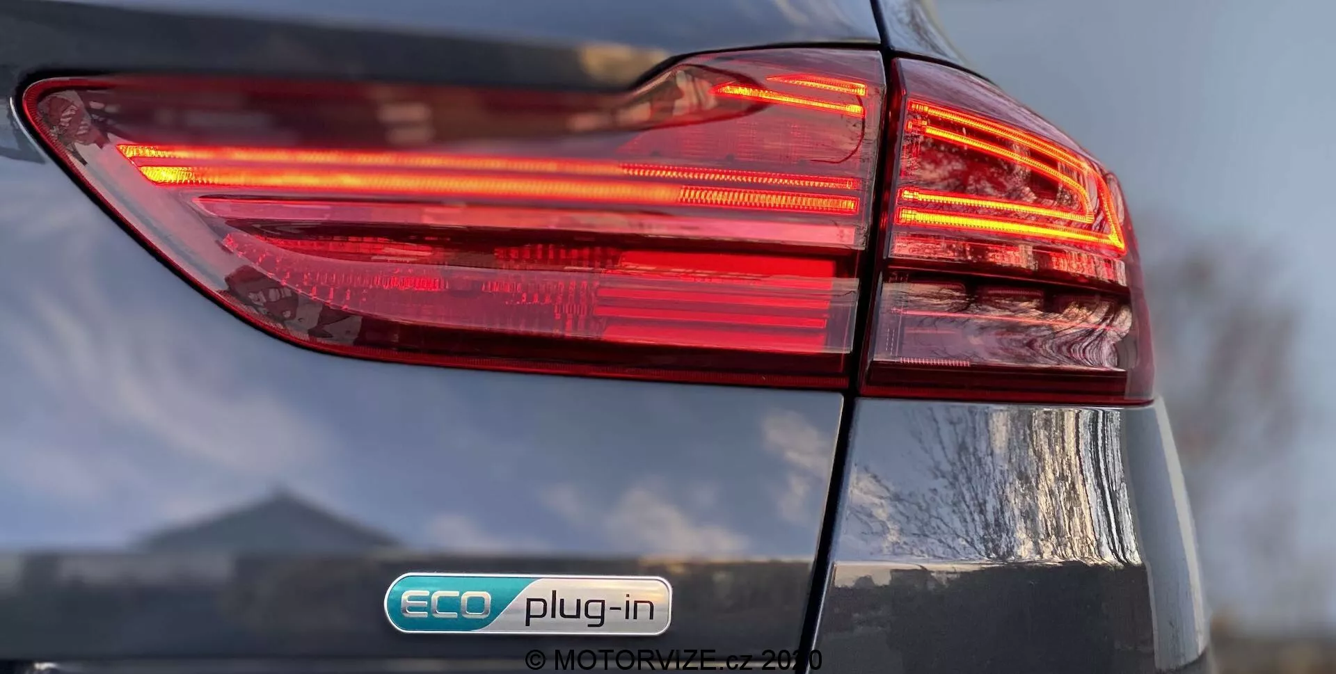 A Kia Ceed 2018 hátsó bal oldalának közeli nézete, amely az illuminált többrétegű kialakítású hátsó lámpacsoportot helyezi előtérbe. Az 'ECO plug-in' jelvény azt jelzi, hogy ez egy plug-in hibrid jármű. A fényes kivitel és a részben látható csomagtérajtó részletessé teszik az autó össztervezését.