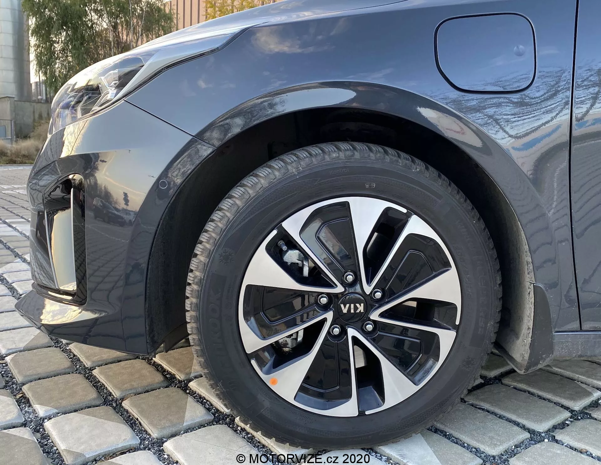 Közelkép alacsony szögből egy 2018-as KIA Ceed jobb első kerékéről, amely többszörösen küllős alu dizájnnal rendelkezik, fekete és ezüst színben, látható futófelület mintázattal, részlegesen látható első sárvédővel, lökhárítóval és a motorháztető egy részével sötét metál árnyalatban.