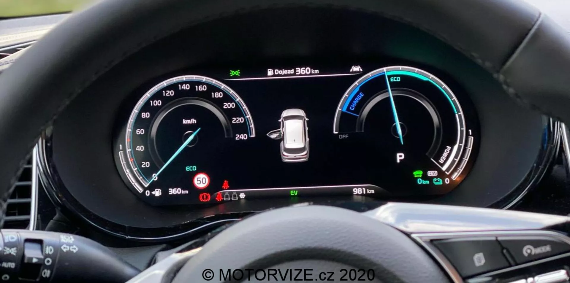 A Kia Ceed 2018 digitális műszerfalának közelképe a vezetői nézőpontból, nagy szélesvásznú kijelzővel és részletes műszerezéssel, beleértve a 260 km/h-ig terjedő sebességmérőt, Eco, Comfort és Power módokkal ellátott teljesítményjelzőt, gumiabroncsnyomás figyelmeztetőt és a 'Park' módjelzőt. A műszerfalon egy 360 fokos kamerarendszer és egy 9811 km-es kilométer-számláló is látható. A környező perem csökkenti a tükröződést, és a kormánykerék gombjai részben láthatók. Vízjel 'MOTORVIZE.cz 2020'.