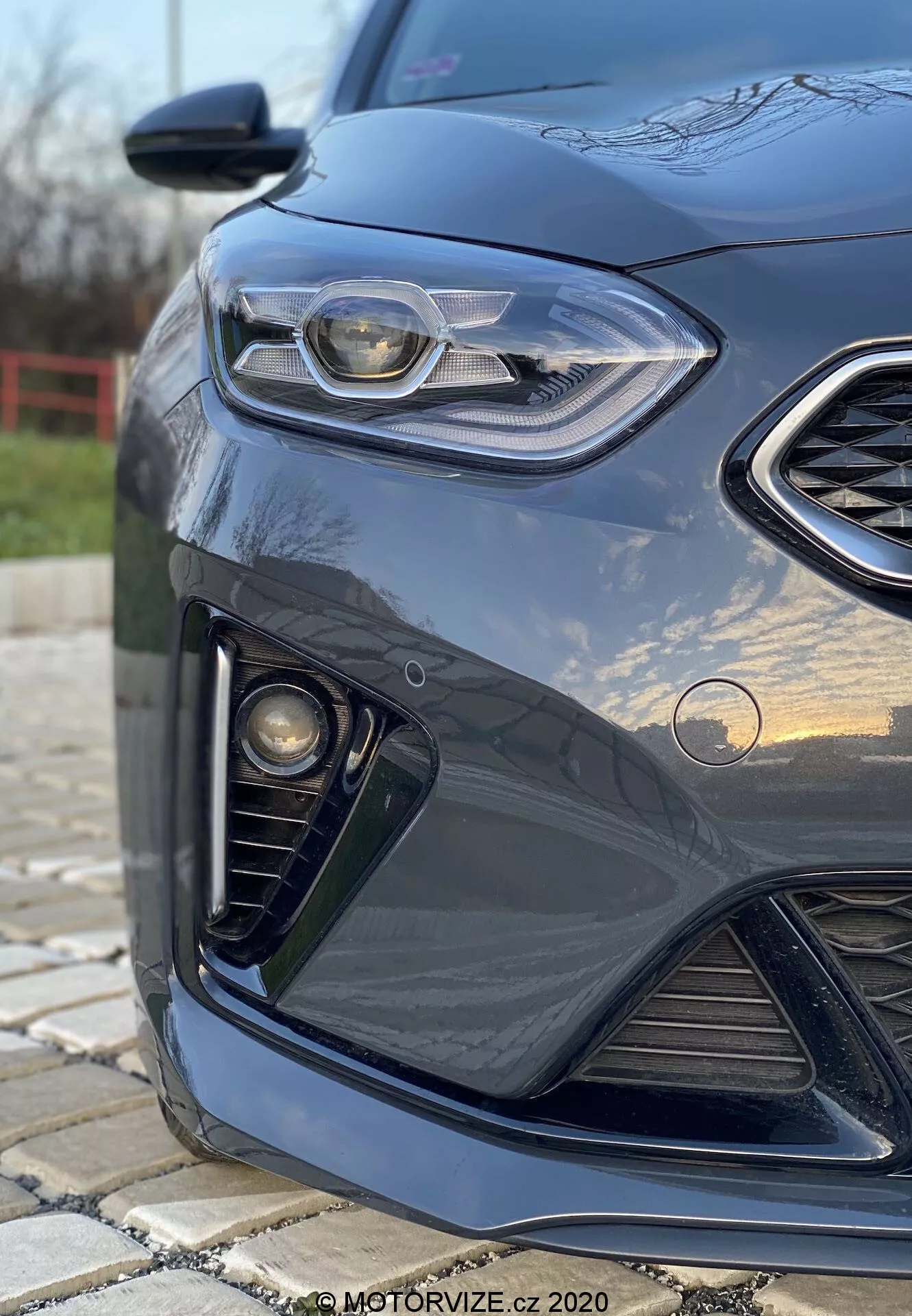 Közeli és szögletes nézet a sötét színű 2018-as Kia Ceed bal első oldaláról, kiemelve a LED-es nappali menetfénnyel ellátott elegáns fényszóró dizájnt, a méhsejtes rácsot, a ködlámpa egységet és az aerodinamikus első lökhárítót, lágy bokeh háttérrel és a fényes festésen lévő tükröződésekkel.