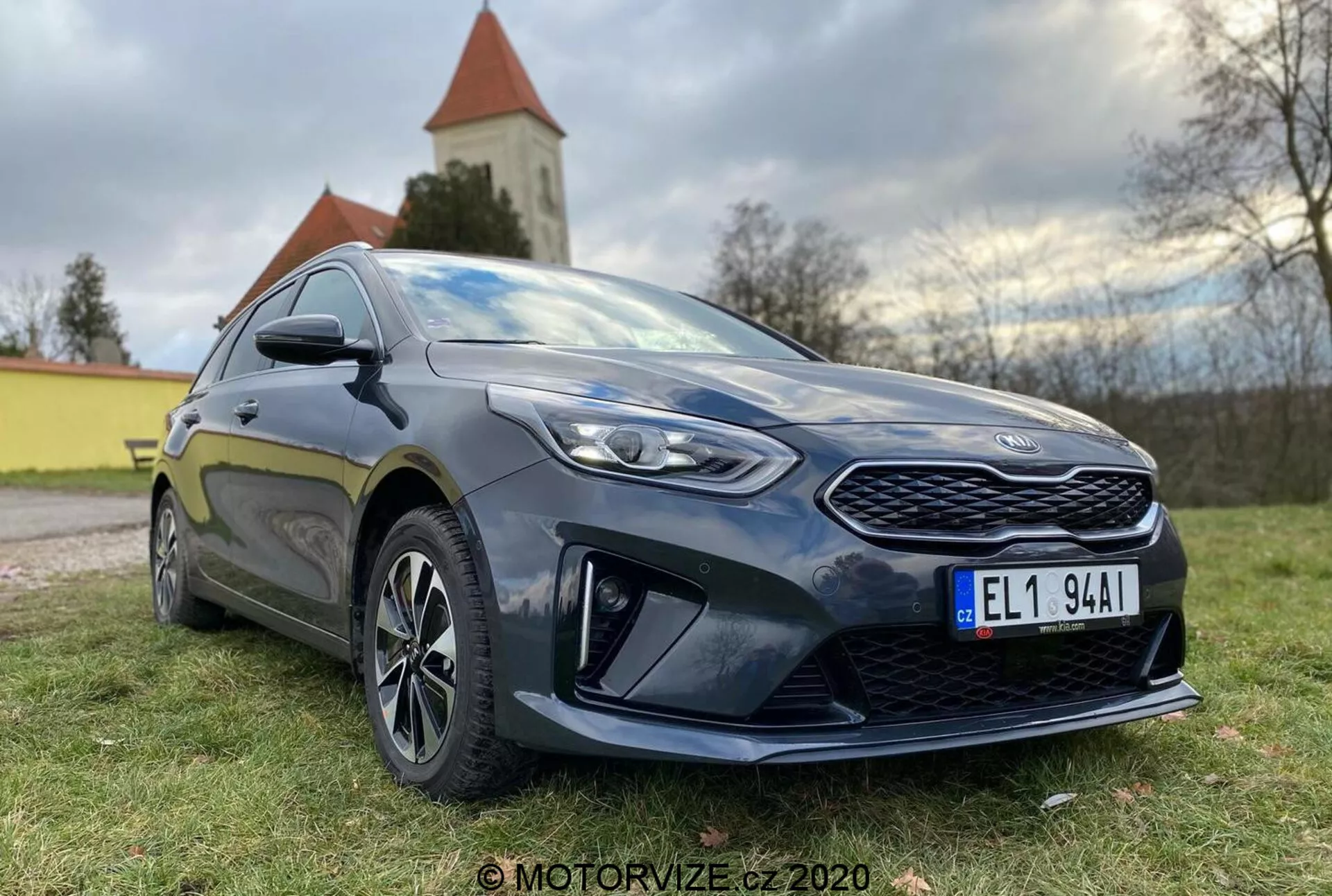 Részletes nézet egy modern Kia Ceed 2018 hatchback autóról, alacsony első háromnegyed szögből fotózva, kiemelve a feltűnő hűtőrácsot, szögletes LED fényszórókat és az oldalsó profilt a vezető ajtajáig. Az autó sötét színű, valószínűleg mélykék vagy fekete, fényes csillogással, ami jól karbantartott állapotát tükrözi. Jelentős dizájnelemek közé tartozik a mintás hűtőrács, éles fényszórók tiszta lencsékkel, az első alsó lökhárítón kifejezett aerodinamikai elemek, többküllős alufelnik, karosszéria színű oldalsó tükrök integrált irányjelzőkkel és sportos oldalsó szoknya. Az autó kiállása kiegyensúlyozott kényelmet és sportosságot sugall. A természetes nappali fény puha megvilágítással kiemeli a részleteket, és a háttérben egy természetes tájkép templommal vagy toronnyal látható.