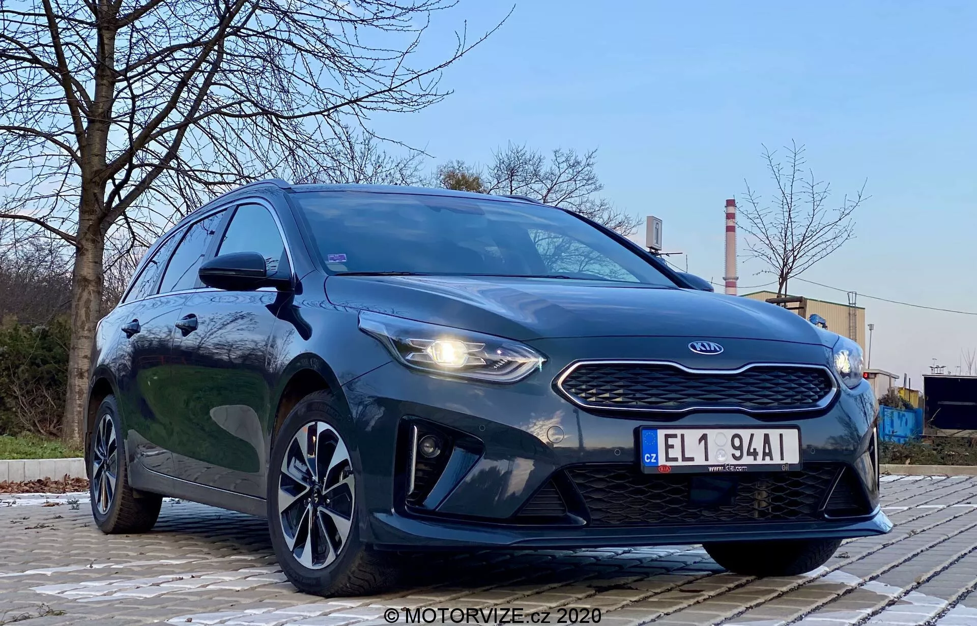 Fotó a 2018-as Kia Ceed-ről alacsony, elülső-bal szögből, kiemelve a jármű hűtőrácsát, fényszóróit és első részletének kialakítását. Az autó agresszív stílusát a feltűnő fényszórók, a tigris orr hűtőrácsa és a motorháztető kontúrjai hangsúlyozzák. A kép LED-es nappali menetfényt, aerodinamikus első lökhárítót és stílusos alufelni kerekeket is láttat. A háttérben fák és ipari struktúrák találhatók, így kontrasztot biztosítanak. A jármű sötét, fémes festése fényes és jól megvilágított, a visszaverődések tiszta felületet jeleznek. A kép az autóra összpontosít alacsony szögű, drámai perspektívában, lágy kültéri megvilágítás mellett.