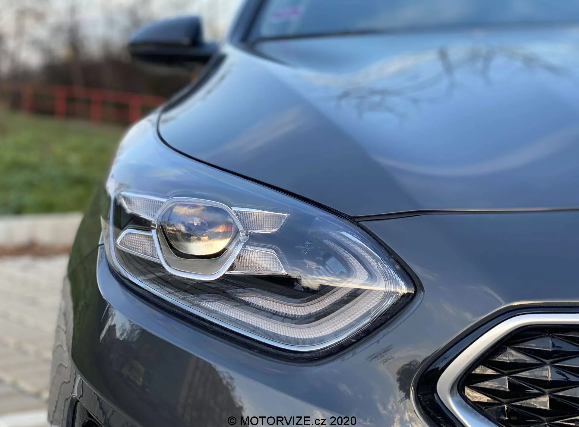 Részletes közelkép a 2018-as Kia Ceed bal első részéről, a kifinomult fényszóróra és az első hűtőrács egy részére összpontosítva. Az alacsony szögből készült felvétel kiemeli az autó elegáns és modern formatervezését, láthatóan fejlett világítástechnológiával, amely magában foglalhatja a LED-es és adaptív fényszórókat is. Az elmosódott háttér kiemeli az autó aerodinamikus elemeit és luxusérzetét, amely teljesítményorientált járművet jelez.