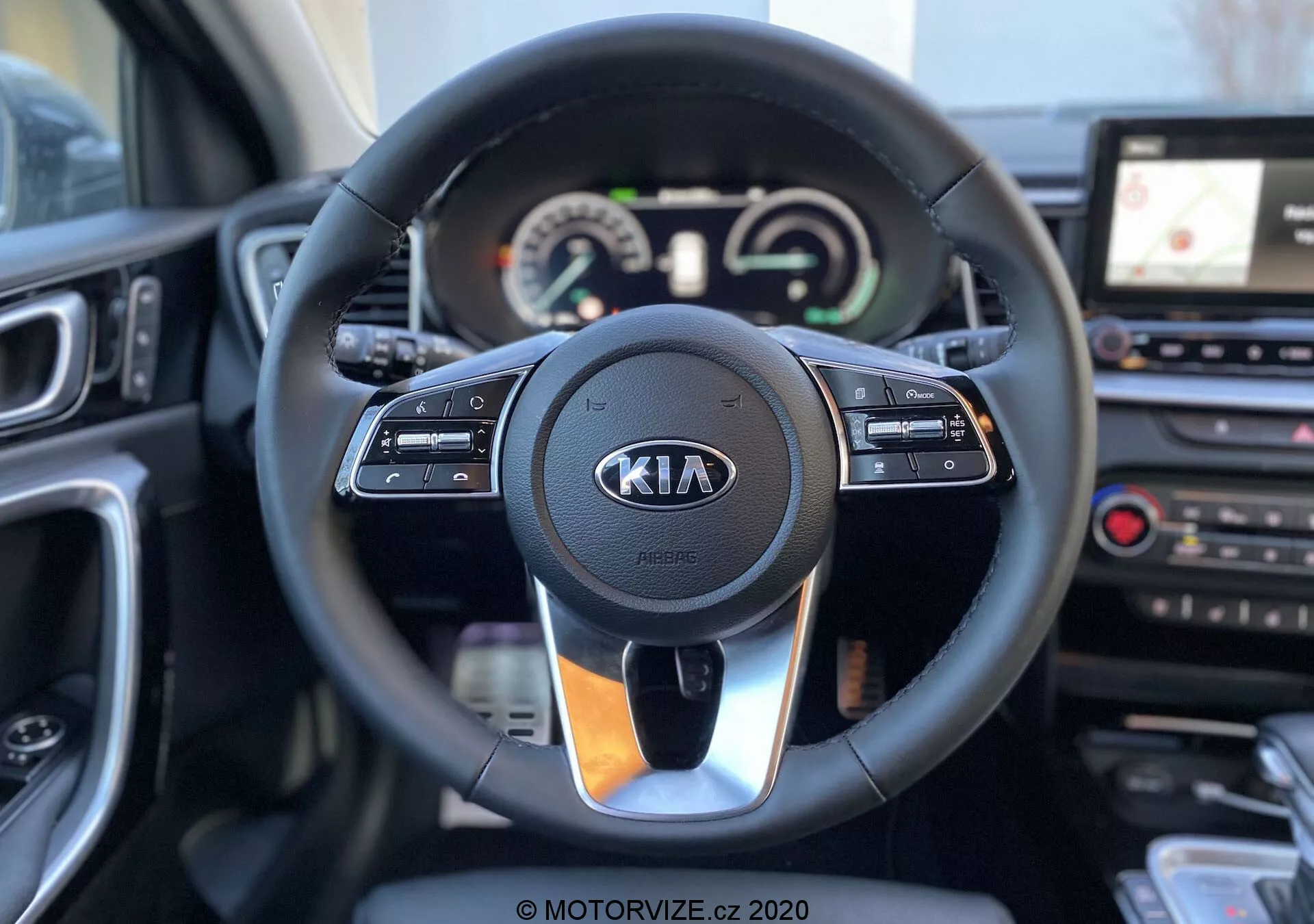 Egy 2018-as Kia Ceed belső nézete a kormánnyal és a műszerfallal a vezetőülés szemszögéből. A modern kormánykerékbe beépített kezelőszervek találhatók a sebességtartó automatika, a multimédiás interakció és a kihangosított kommunikáció számára, jól látható Kia logóval. A kormány mögött helyezkedik el a műszerfal, amely analóg tárcsákat és digitális kijelzőt kombinál; ezek a sebességmérőt és a fordulatszámmérőt, valamint egy többfunkciós kijelzőt mutatnak. A kormánykerék jobb oldalán látható a középső konzol egy része, amely érintőképernyős infotainment rendszert és GPS-navigációt, valamint klíma vezérlő kapcsolókat tartalmaz. A kép tiszta és éles, sekély mélységélességgel, amely kiemeli a kormánykereket és annak közvetlen környezetét.