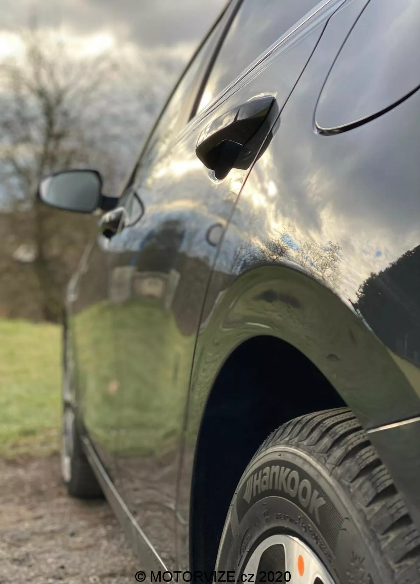 Közelről készült oldalkép a 2018-as Kia Ceed-ről, kiemelve a hátsó kereket 'Hankook' gumival, az áramvonalas ajtókilincset és a részben elmosódott oldalsó tükröt. A kép alacsony szögből készült, sekély mélységélességgel, ami élesen fókuszál a hátsó kerékre és az ajtó alsó részére, miközben a háttér művészien elmosódott. Az autónak sötét, fényes felülete van, mely tükrözi az eget és a környezetet, kiemelve az elegáns dizájnvonalakat és a jól karbantartott karosszériát.