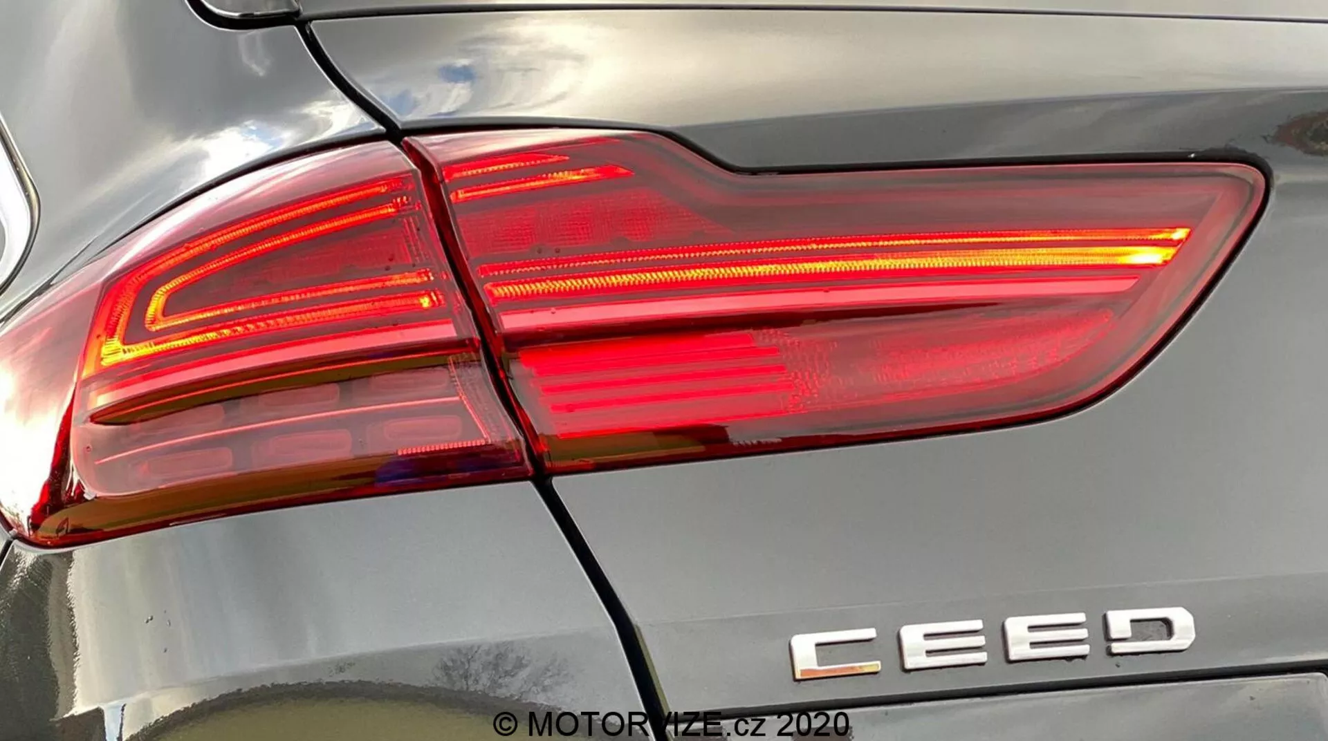 Közelről részletes nézet egy 2018-as Kia Ceed hátsó jobb oldali lámpatestéről, amely világít, és modern, éles, szögletes vonalakkal, piros és átlátszó lencseszekciókkal, és 'CEED' modell jelvénnyel rendelkezik. Egy vízszintes vonal elválasztja a hátsó lámpát a hátsó lökhárítótól. Az autó karosszériáján lévő tükröződések kiemelik a felület görbülését. 'MOTOR1.cz ©2020' vízjel.