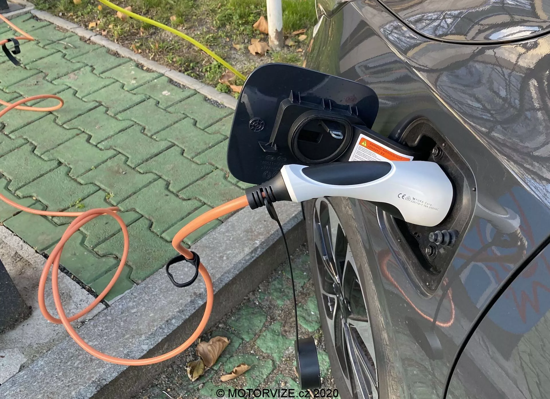 Közeli oldalnézet egy szürke elektromos járműről (EV) töltés közben, kiemelve a nyitott töltőportot és a járműhöz csatlakoztatott narancssárga EVSE csatlakozót. A modern fehér és fekete töltőcsatlakozó LED-jelzőfénnyel rendelkezik. A kép a töltési folyamatra összpontosít, kültéri burkolt területen, szétszórt levelekkel.