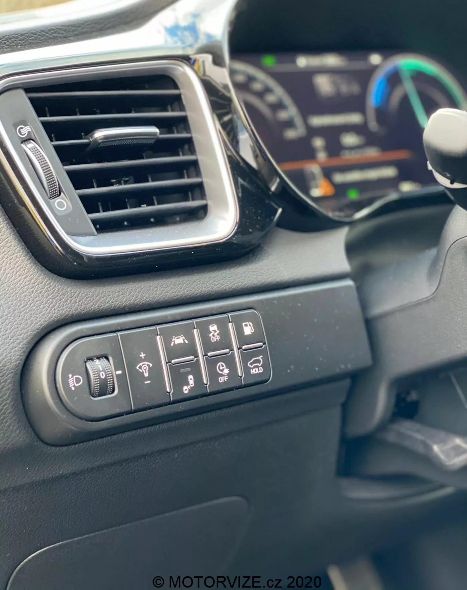 A Kia Ceed 2018 belső terének közeli képe, amelyen a külső tükrök állítására, a hátsó ablak fűtésére, a kormánykerék fűtésére, a sávelhagyásra figyelmeztető rendszerre, a parkolássegítő rendszerekre és a vonóerő-szabályozásra szolgáló vezérlőgombok láthatók. A képen továbbá egy elegáns szellőzőnyílás, a homályos háttérben egy digitális műszercsoport részlete és a kormánykerék széle látható. A belső anyagok többnyire kemény műanyagból készültek matt felülettel, nappali fényben megörökítve.