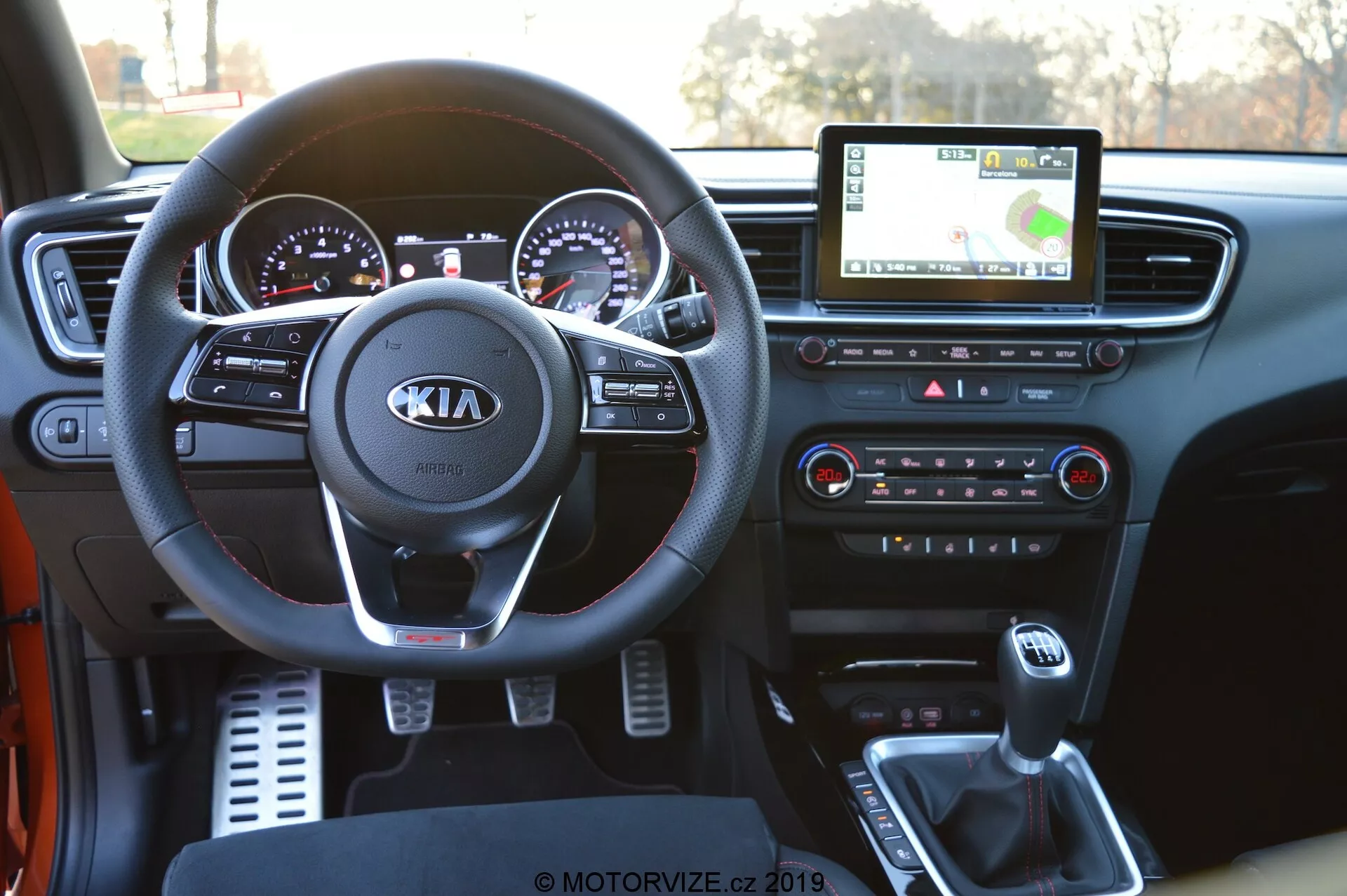 A 2021-es Kia Ceed belső nézete a vezető perspektívájából, amely bemutatja a részletes műszerfalat. A képen kiemelkedően látható a Kia logóval ellátott kormánykerék, valamint az audió-, hívás- és sebességtartás-szabályozó gombok. Mögötte a krómozott analóg műszereket és egy digitális kijelzőt tartalmazó műszercsoport látható. A központi infotainment érintőképernyője GPS navigációt jelenít meg, alatta pedig a klímavezérlő gombok és tekerők találhatók. A középkonzolon a váltókar, pohártartók és egy USB port látható, amely a modern autós technológiát hangsúlyozza.