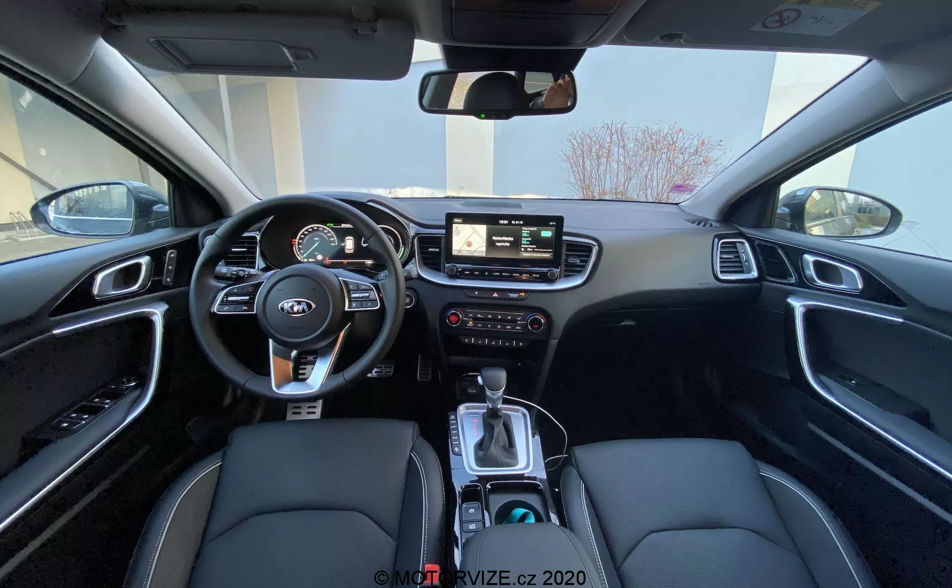 Széles látószögű nézet a 2018-as Kia Ceed belső teréről a hátsó ülésről, amely bemutatja a vezető területét, beleértve a kormánykereket beépített vezérlőkkel, a digitális és analóg műszerekből álló műszerfalat, a központi infotainment rendszert érintőképernyővel, az infotainment és klímarendszer kézi vezérlőelemeit, valamint a középkonzolt váltókarral, vezetési módválasztóval, pohártartókkal és fekete belső teret, szintetikus bőr ülésekkel, fehér varrással és csiszolt alumínium díszítéssel.