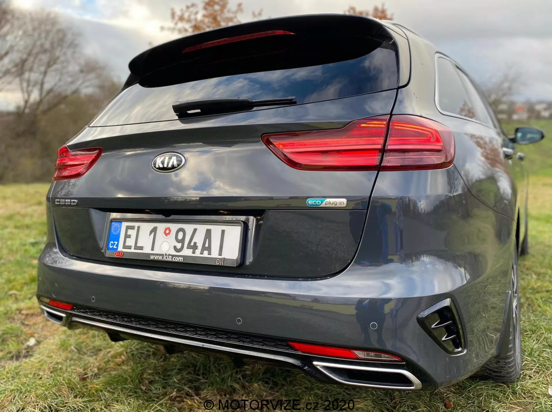 Egy sötétszürke 2018-as Kia Ceed Proceed modell hátulnézete, részletes jellemzőkkel, mint a vízszintesen orientált LED hátsó lámpák, tetőre szerelt hátsó spoiler, dupla krómozott kipufogóvégek, fekete diffúzor stílusú hátsó lökhárító és központi emblémák, köztük 'Kia' és 'GT Line'. A központilag elhelyezett rendszámtábla feletti tolatókamera parkolási asszisztensre utal, míg a sekély mélységélesség kiemeli az autó márkáját és modellazonosítóit a füves háttér előtt.