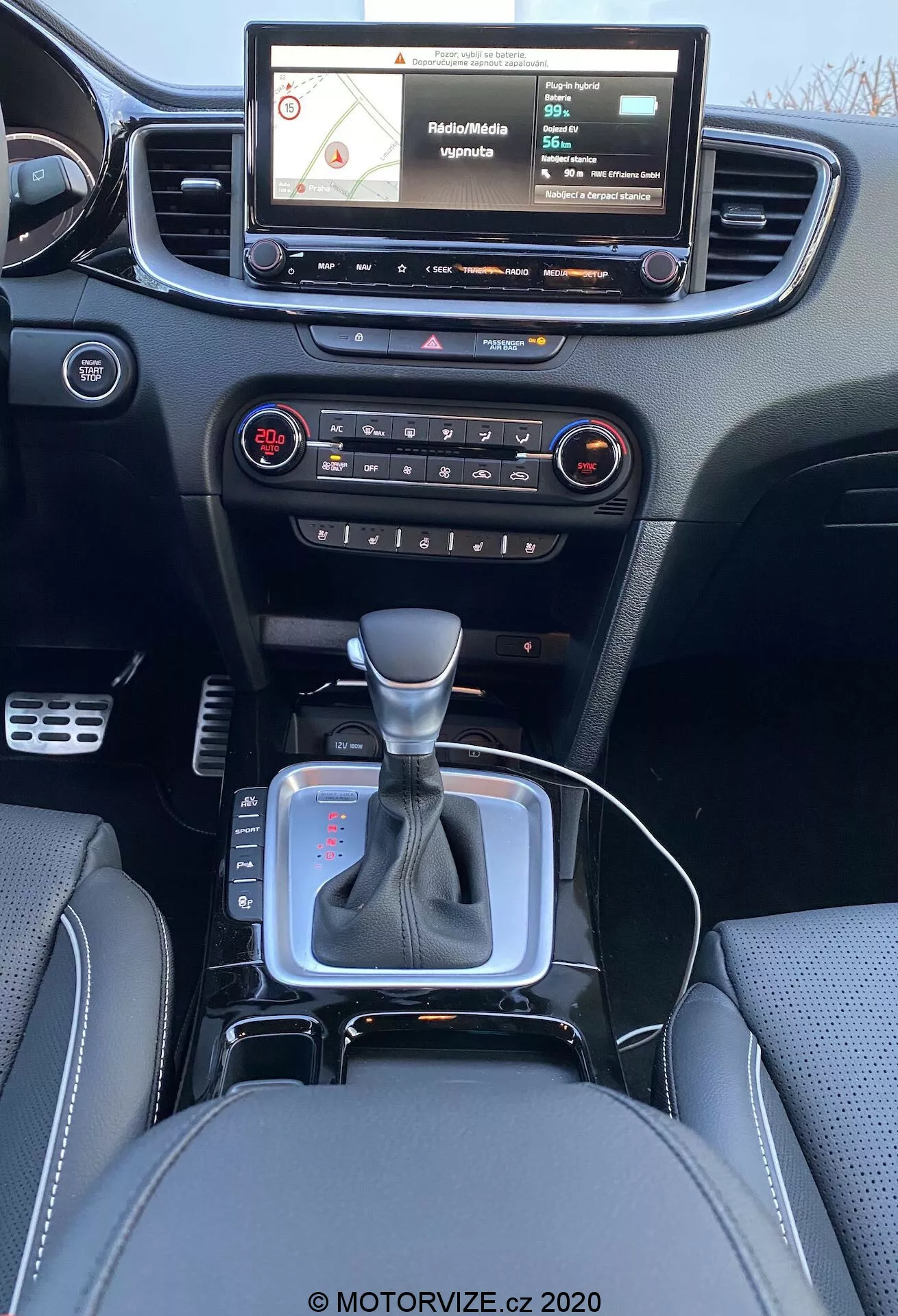 2018-as Kia Ceed belső nézete a hátsó ülés perspektívájából, amely egy modern műszerfalat mutat be érintőképernyős infotainment rendszerrel, ami navigációs térképet jelenít meg, fizikai gombokkal és vezérlőkkel az audio és klímaberendezés vezérléséhez, központi szellőzőnyílás, semleges helyzetben lévő váltókar ezüst díszítéssel és fekete bőr borítással, kis tárolóhely a váltókar közelében, valószínűleg 12V-os aljzathoz vagy USB portokhoz, bőr ülések részletes varrással.