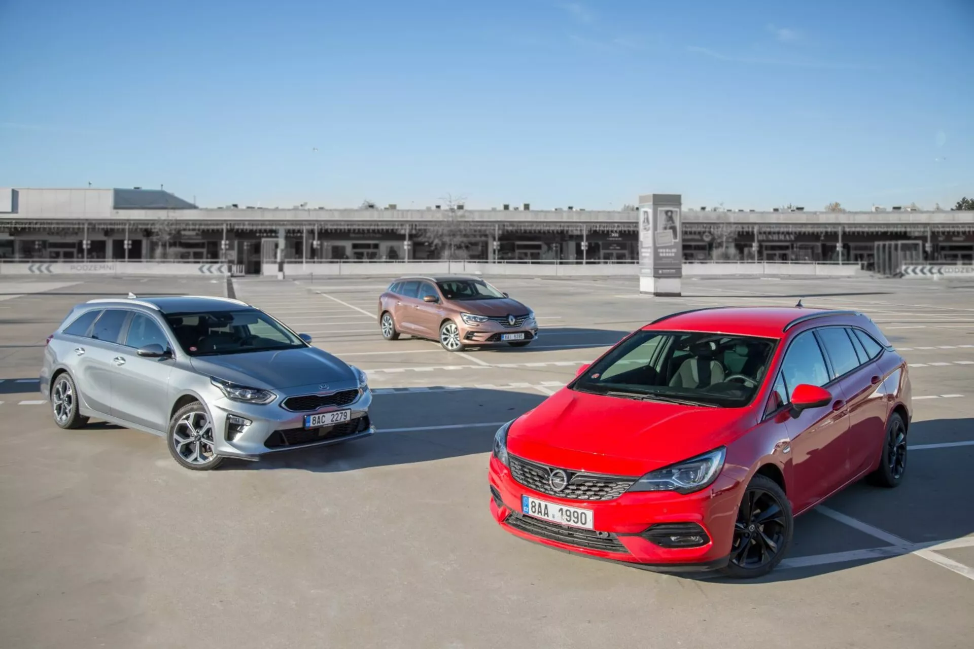 Három Kia Ceed modell 2009-ből, beleértve egy piros ferdehátút, arany crossover-t és ezüst kombit, egy tetőtéri parkolóban parkolva, magasból fotózva, bemutatva az autók elejének dizájnját, oldalnézeteit és hosszúságát