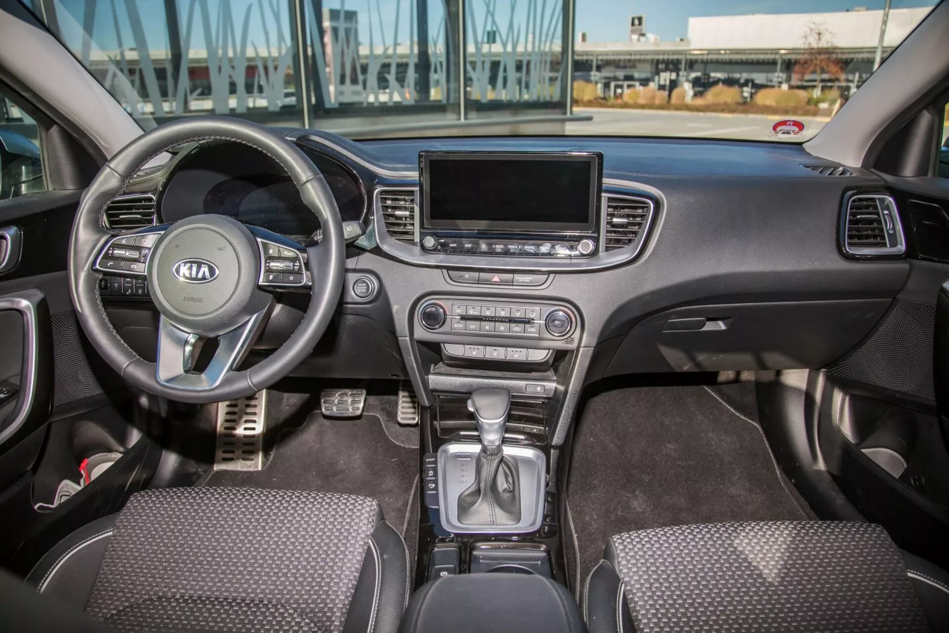 2009-es Kia Cee'd belső terének megjelenítése az első kabin teljes területével, beleértve a multifunkciós kormánykereket Kia logóval, a kormány mögötti műszerfal részt, a park pozícióban lévő váltókart a középkonzolon, a klímavezérlő gombokat, az infotainment képernyőt, a szövetborítású üléseket és a vezető és utas oldali ajtóvezérlőket természetes nappali fényben