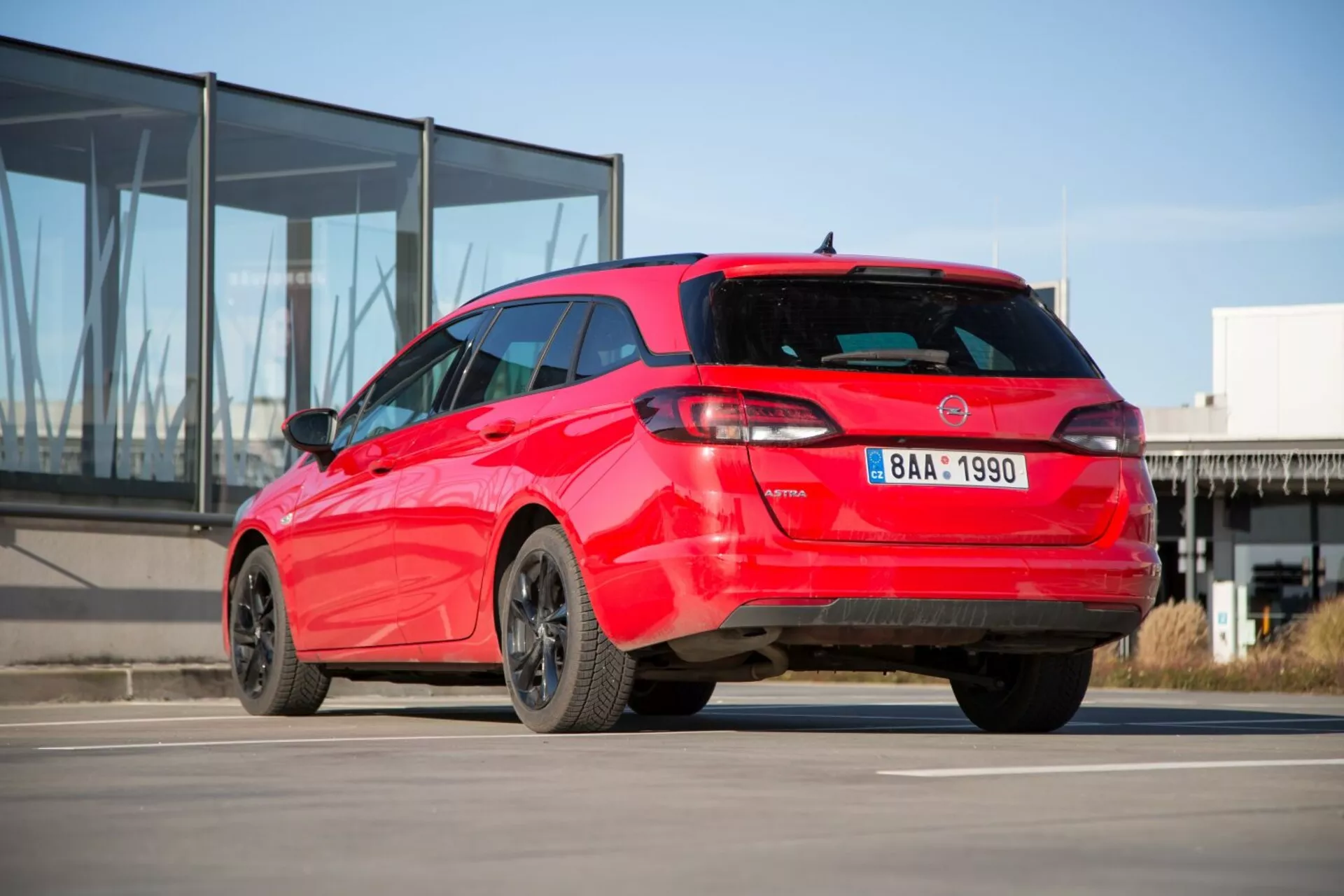 2009-es piros Kia Ceed hatchback sportos háromnegyedes hátsó nézetén betonon modern üveg homlokzatú épület hátterével, különleges hátsó lámpa kialakítással, tetős spoilerrel, cápauszony-antennával, ötvözött felnikkel és lehetséges parkolássegítő szenzorokkal.