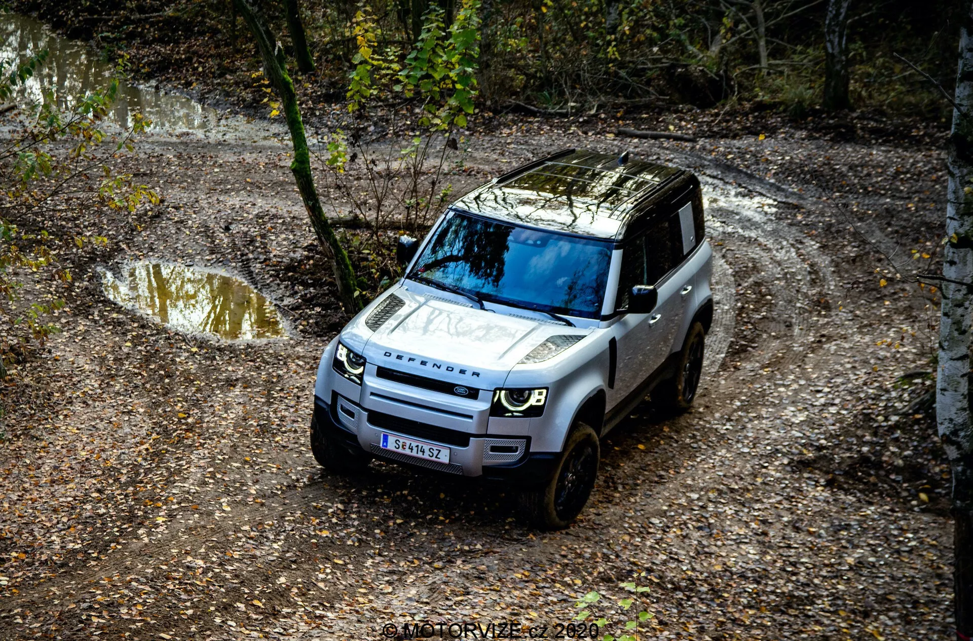 Madártávlatból készült kép egy 2020-as Land Rover Defenderről, amely egy terepjáró ösvényen halad, és jól láthatóak rajta az első rács, a motorháztető, a szélvédő és a vezető oldalának egy része. Az útvonalon víz- és sárrészek találhatók, amelyeket fák vesznek körül.