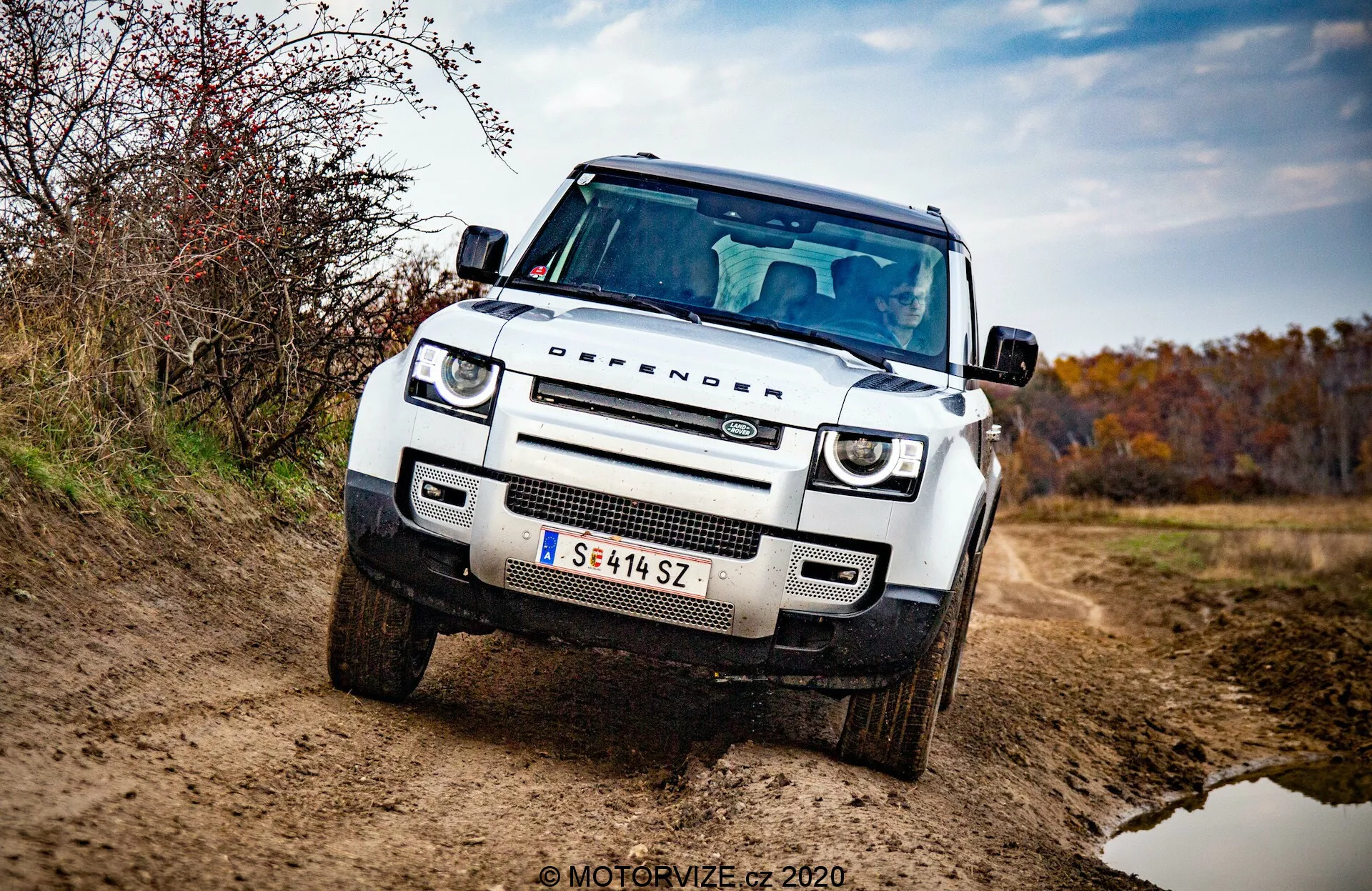 2020-as Land Rover Defender elölnézet háromnegyed szögből, fehér, szögletes terepjáró lapos motorháztetővel, szögletes kerékjárati ívekkel, kör alakú LED nappali menetfénnyel, adaptív fényszórókkal, méhsejt rácsos hűtőráccsal és márka logóval, elülső lökhárító védőlemezzel, megemelt és robusztus állásban, oldalsó tükör beépített irányjelzőkkel és hangsúlyozva a hűtőrácsot, fényszórókat és első lökhárítót. Jármű kavicsos úton, külső részén szennyeződéssel, földes, őszi környezetben. Professzionális, jól megvilágított fénykép elmosódott háttérrel és 'MOTORVIZE © 2020' vízjellel.