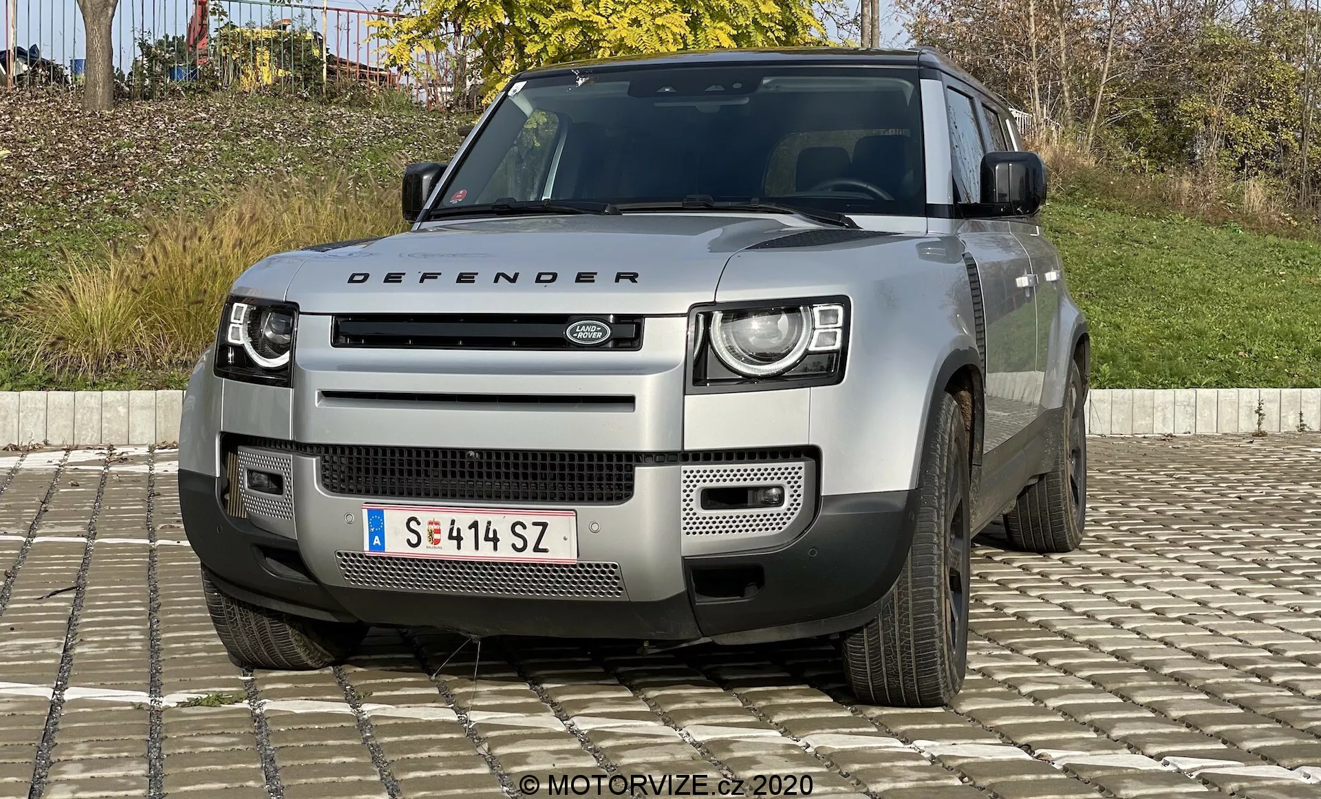 A Land Rover Defender 2020-as modell elölnézeti képe, amely kiemeli jellegzetes kétszintes LED fényszóróit, a Land Rover logóval ellátott kiemelkedő hűtőrácsát, geometriai lökhárítóját ezüst védőlemezzel és egyéb robusztus tulajdonságait, amelyek off-road képességekre utalnak. Látható még a motorháztető a 'Defender' névtáblával, a légbeömlők és az ötágú kerekek részleges nézete is. A jármű világos, semleges színű, valószínűleg ezüst vagy szürke.