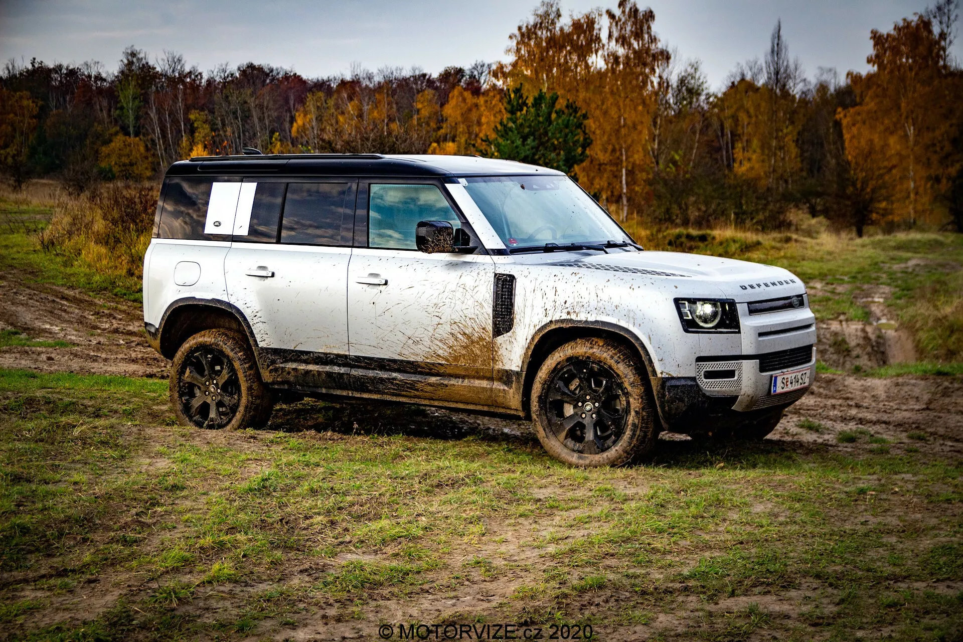 Oldalnézet egy Land Rover Defender 2020 terepjáróról háromnegyedes szögből, fehér karosszériával, fekete tetővel és kerekekkel, sárral borítva. A jármű átlósan elhelyezkedik a kép bal alsó sarkától a jobb felső sarokig, sáros, természetes terepen, borongós ég alatt, őszi fákkal a háttérben.