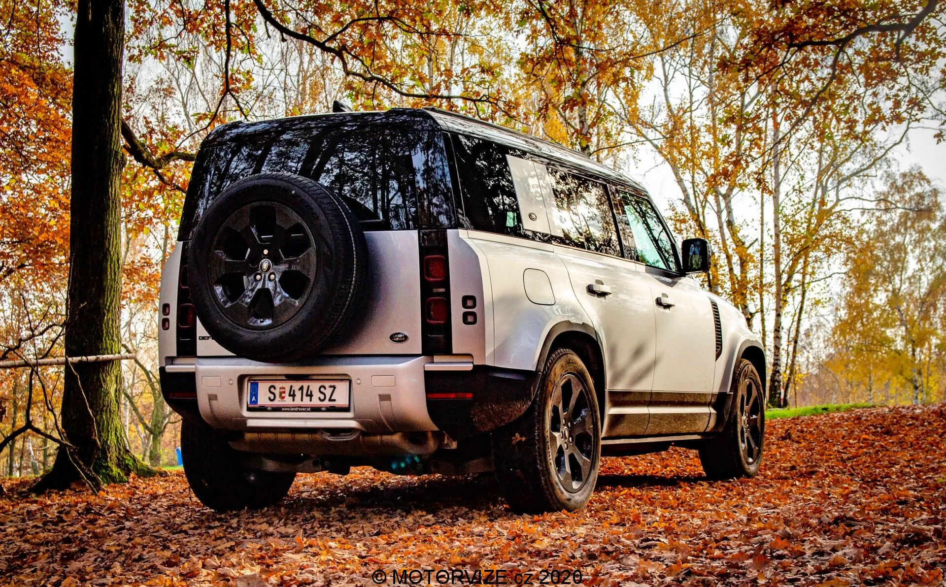 Háromnegyedes hátsó nézet egy modern SUV-ról, amely egy őszi erdei ösvényen áll, levelekkel borított, kiemelve a jármű magasságát és talajszabadságát terepjáró képességeinek bemutatására. A felvételen látható a hátsó rész, beleértve a hátsó ajtót, a tető egy részét, a hátsó ablakot és az egyik oldalsó profilt. Látható jellemzők: a függőleges hátsó lámpa kialakítás, robusztus fekete ötvözetű kerék vastag futófelülettel, a hátsó ajtóra szerelt pótkerék, a hátsó lökhárító parkolószenzorokkal, kéttónusú festés kontrasztos tetővel, hátsó ablaktörlő, a rendszámtábla alatt található tolatókamera, és az oldalsó lépcsők a kabinba való könnyebb bejutáshoz. A szögletes, robusztus karosszéria luxust sugall, a markáns karaktersor vonallal az oldalsó ajtók mentén. A természetes, szórt fény felhős napot vagy fák kertinakápolásán keresztül szűrődő fényt sugall, fokozva a szezonális, szabadtéri témákat.