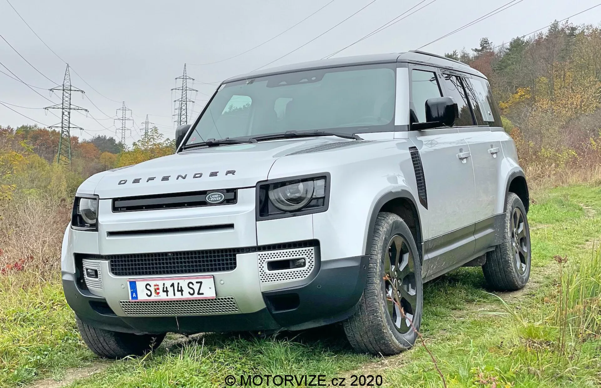 Ez a fénykép egy modern Land Rover Defender 2020 SUV-t ábrázol elülső negyedik szögből, bemutatva a vezető oldalát. A jármű enyhén terepen, füves területen helyezkedik el, egy földút nyomaival, ami kiemeli terepjáró képességeit. A jellegzetes dobozos formatervezés nagy hasmagassággal és két tónusú színsémával rendelkezik: fehér karosszériával és ellentétes fekete tetővel. Az elülső rész retro ihletésű LED fényszórókat, Defender emblémával rendelkező motorháztetőt és minimalista elülső rácsot mutat, kiemelve a terepen való megbízhatóságát. A látható technikai jellemzők közé tartoznak az első lökhárítóba integrált parkolásérzékelők, amelyek fejlett vezetéstámogató rendszerekre utalnak. Az erős kerékívek nagy terepjáró abroncsokat fogadnak be, és a tervezési elemek, például az alufelnik és az egyedi rács dizájnja hangsúlyozottak.