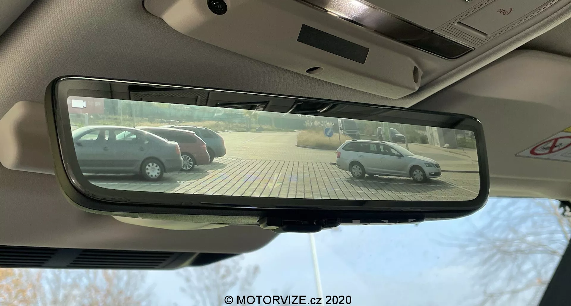 Kilátás egy 2020-as Land Rover Defender sofőrüléséből, a belső téglalap alakú, automatikus fényerő-szabályozású visszapillantó tükörre fókuszálva, amely két ezüstszínű ferdehátú autót tükröz vissza, amelyek merőlegesen parkolnak egy bézs burkolólapokból készült parkolóban, napsütéses napon.