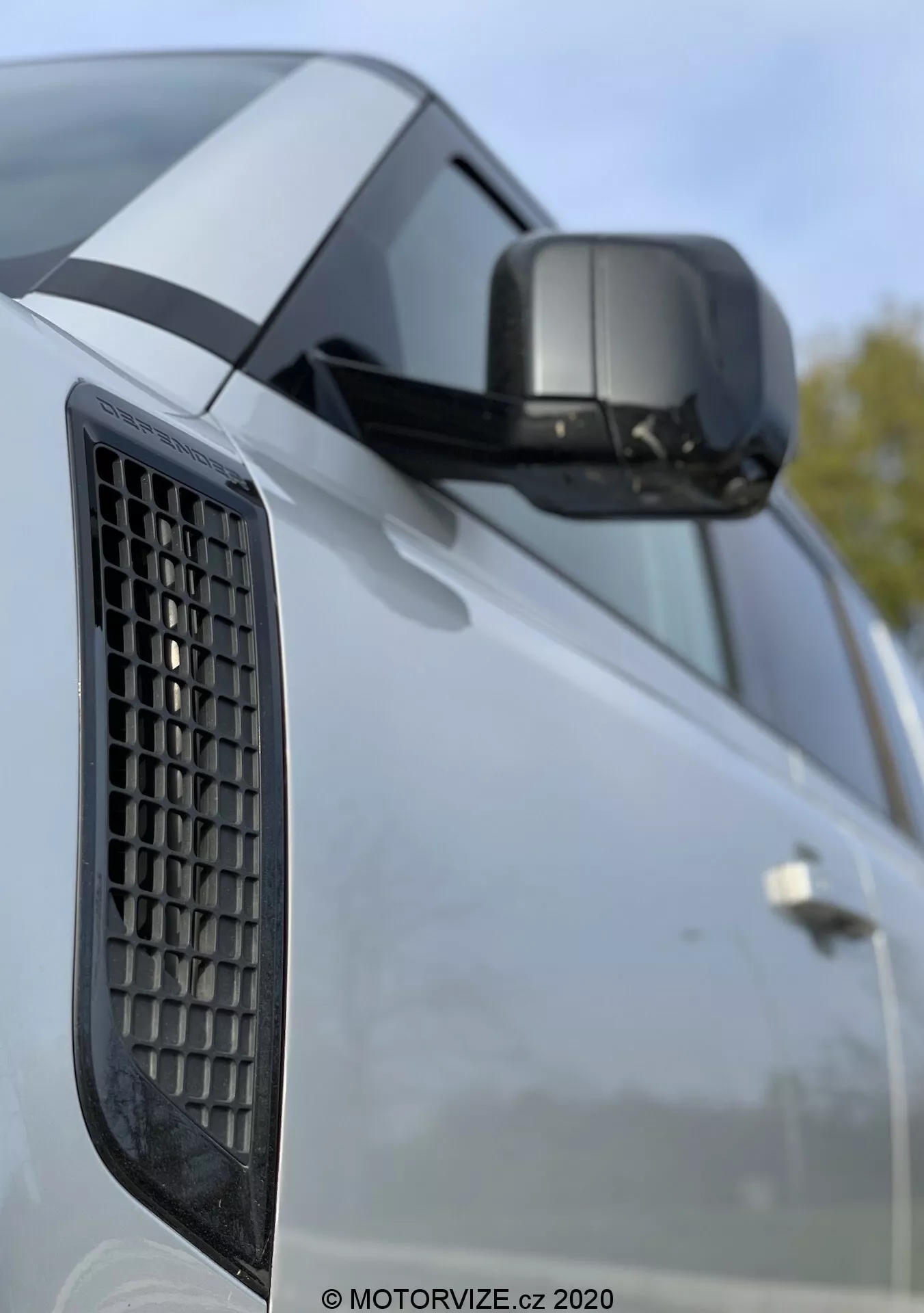 2020-as Land Rover Defender közelről, amely az oldalsó sárvédőt rácsos szellőzővel, az integrált irányjelzővel ellátott oldalsó tükröt és az ajtó részét kiemeli éles karaktervonalakkal; a kép a jármű formatervezési elemeire összpontosít, homályos háttérrel, amely kültéri környezetet javasol.