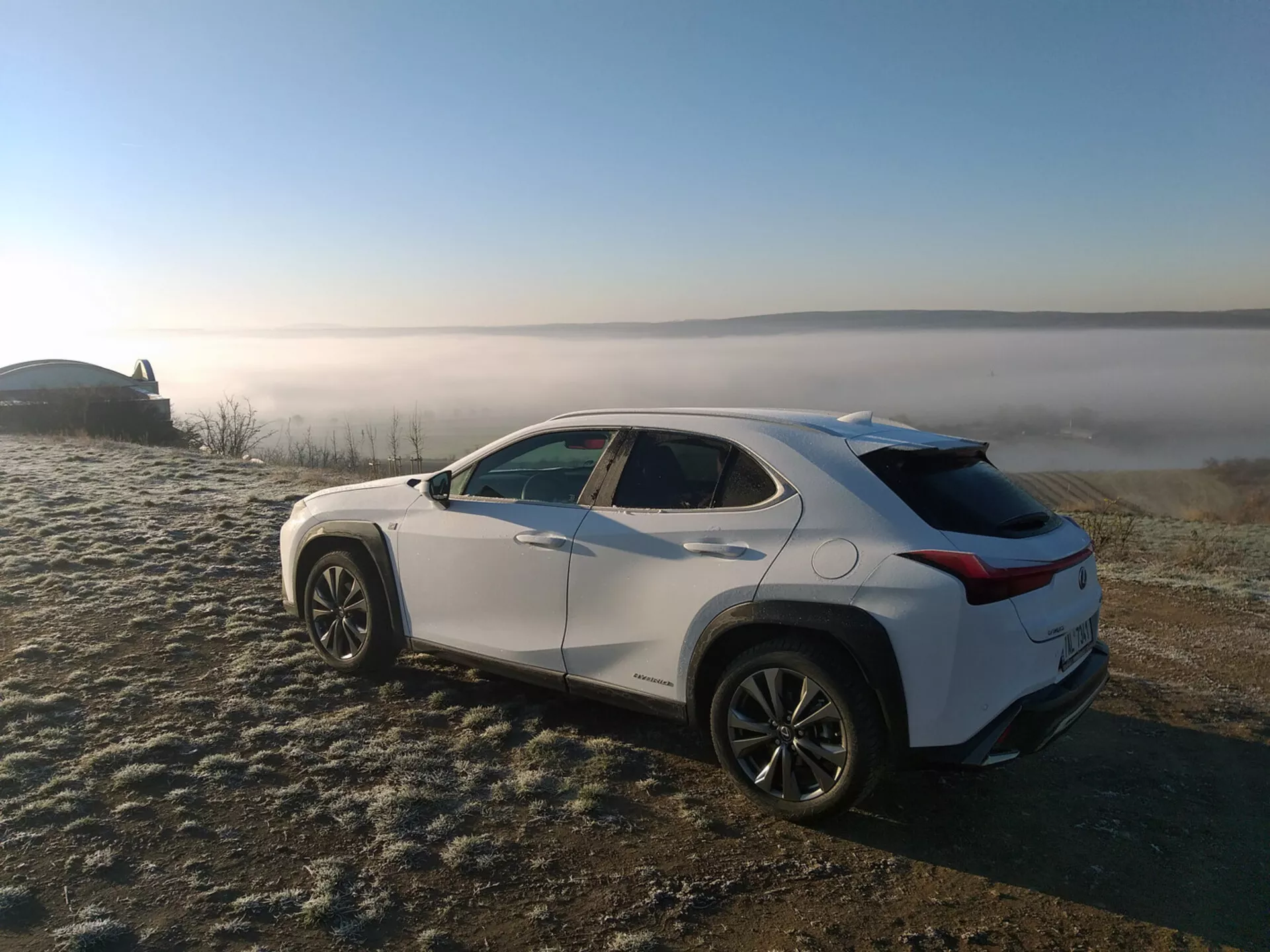 2019-es Lexus UX crossover SUV, fagy által borított füvön parkolva, ködös tájkép hátterével, bemutatva a jármű profilját egy hátsó háromnegyed szögű nézetből, kiemelve az oldalsó karaktervonalakat, kerékíveket, fokozatosan vékonyodó tetővonalt és ötvözött kerekeket.