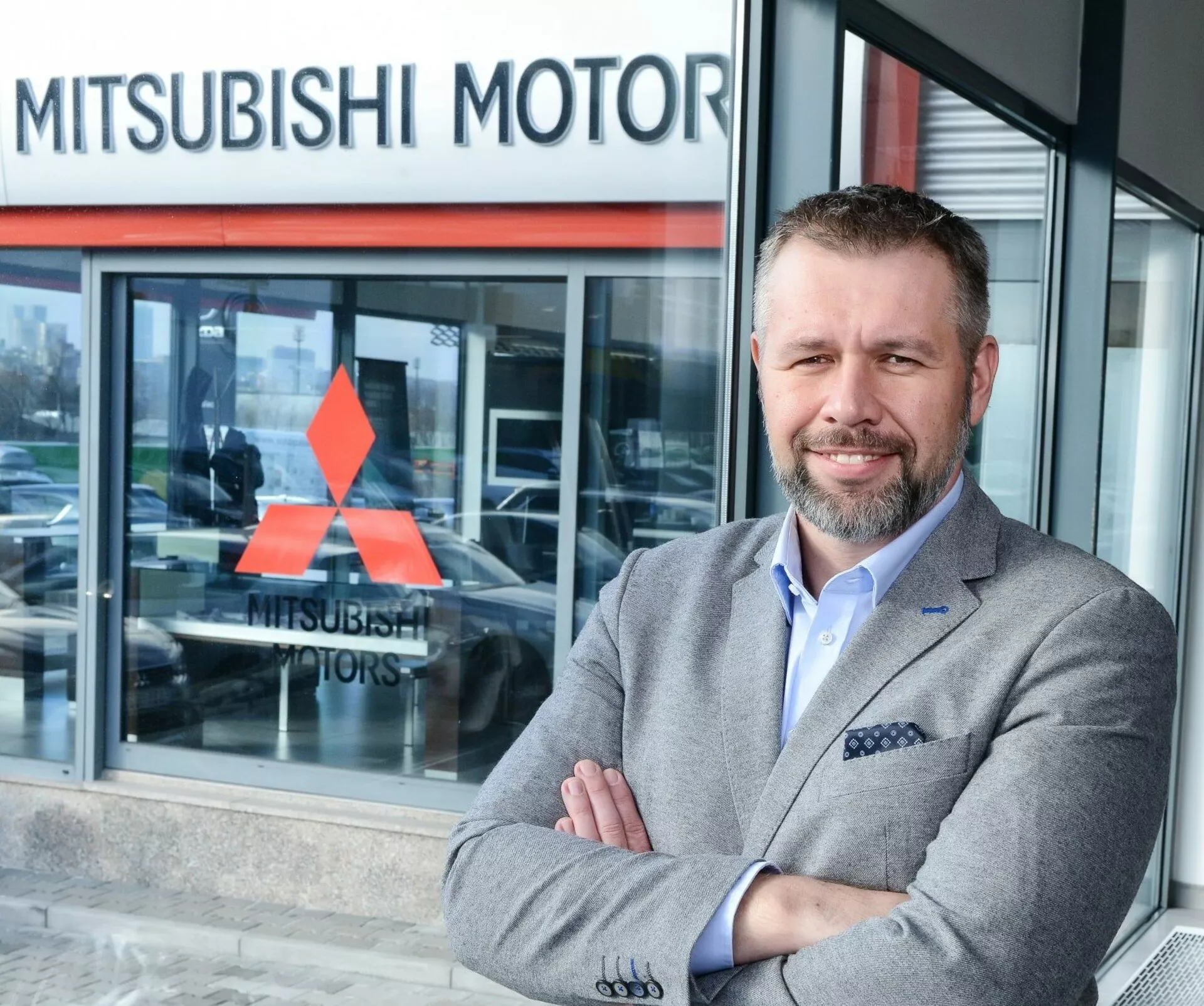 Karba tett kézzel álló személy a Mitsubishi Motors autószalon előtt, részben látható jármű az üvegablakon keresztül, ami a háttérben egy kiállítási modellt sugall