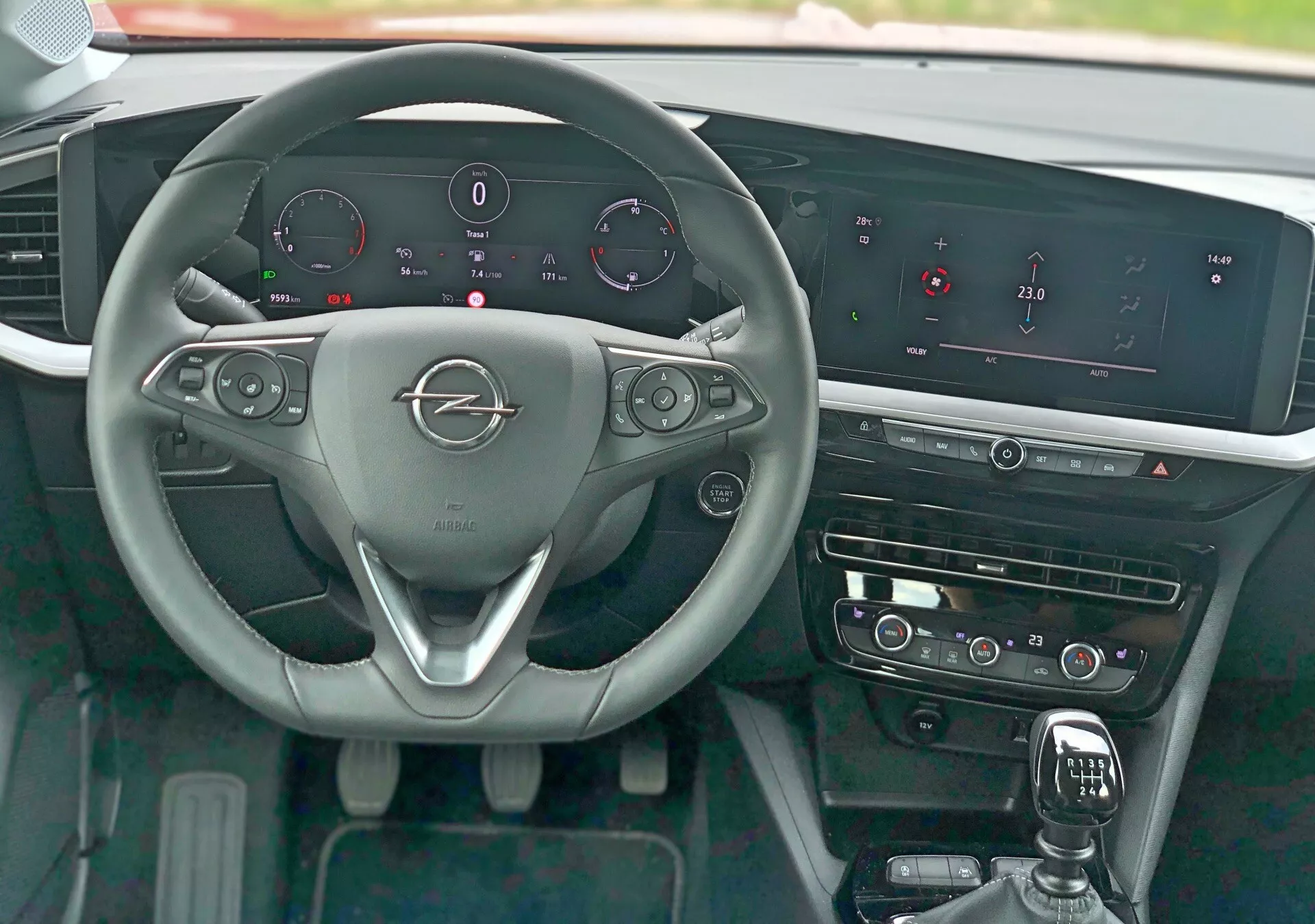 A 2021-es Opel Mokka belső nézete a vezetőülés perspektívájából, amely megmutatja a háromküllős kormánykereket fém márka-emblémával, kormánykerékbe épített vezérlőkkel az audió, telefon és tempomat funkciókhoz, váltófülekkel, digitális műszercsoporttal, amely a sebességet, a fordulatszámot, az üzemanyagszintet és a motor hőmérsékletét mutatja, kiemelkedő infotainment rendszer széles vízszintes képernyővel, érintésérzékeny HVAC vezérlőkkel, bőr borítású váltókarral, polírozott díszítőelemekkel és kulcs nélküli indítógombbal.