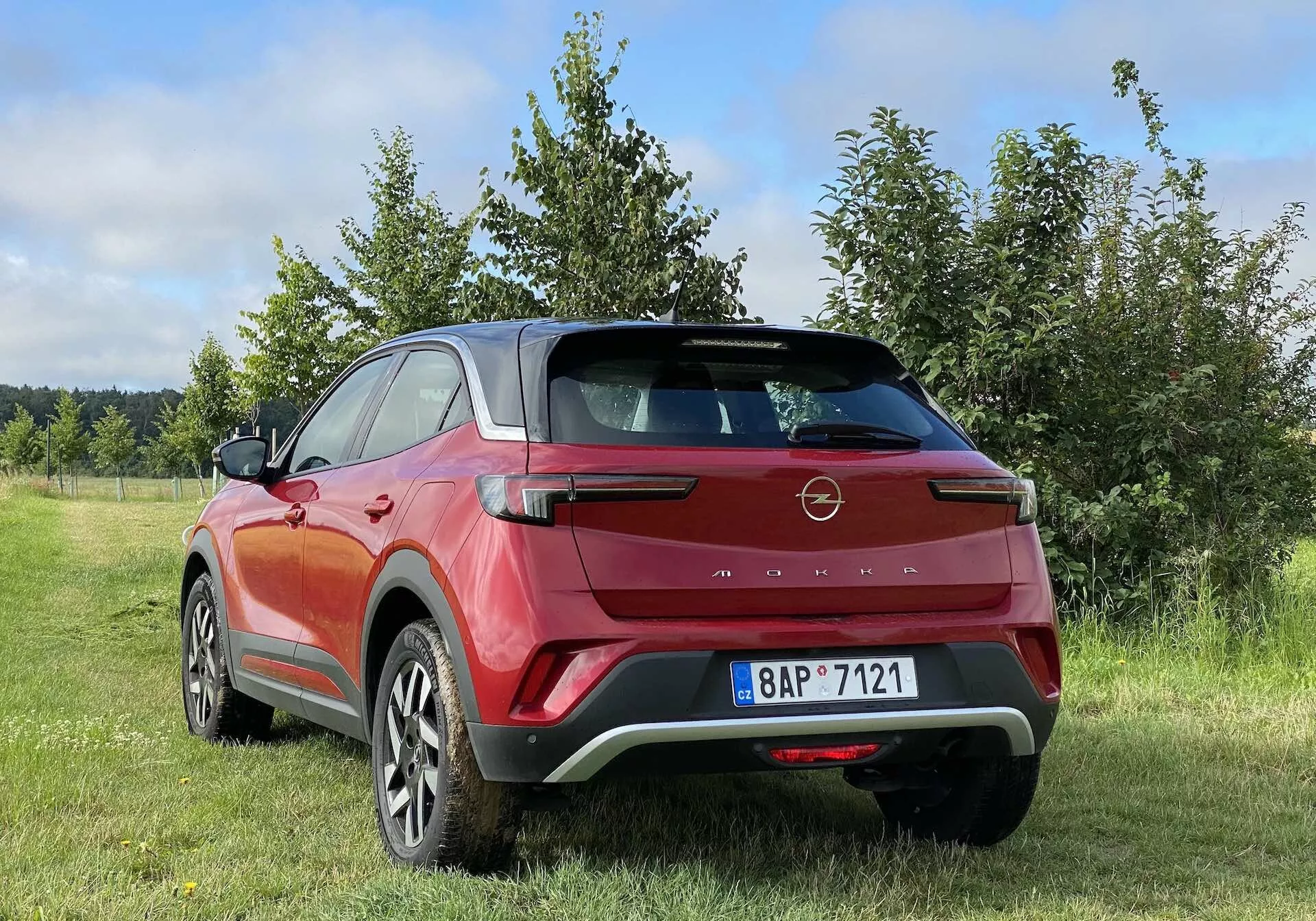 Színes fénykép, amely egy Opel Mokka 2021-es modern crossover SUV hátsó háromnegyedes nézetét ábrázolja piros-fekete színben, egy füves területen parkolva, a háttérben fákkal vagy bokrokkal. A jármű dizájnelemei közé tartozik az aerodinamikai hátsó spoiler, a szögletes LED hátsó lámpák, a magasan elhelyezett féklámpa, a két tónusú karosszéria, a nagy könnyűfém kerekek, a süllyesztett ajtókilincsek és a fekete műanyag burkolat. A fotó alacsony szögből készült, kiemelve az SUV magasságát és távolságát a talajtól, részben felhős égbolttal, amely változékony időjárási körülményeket mutat.