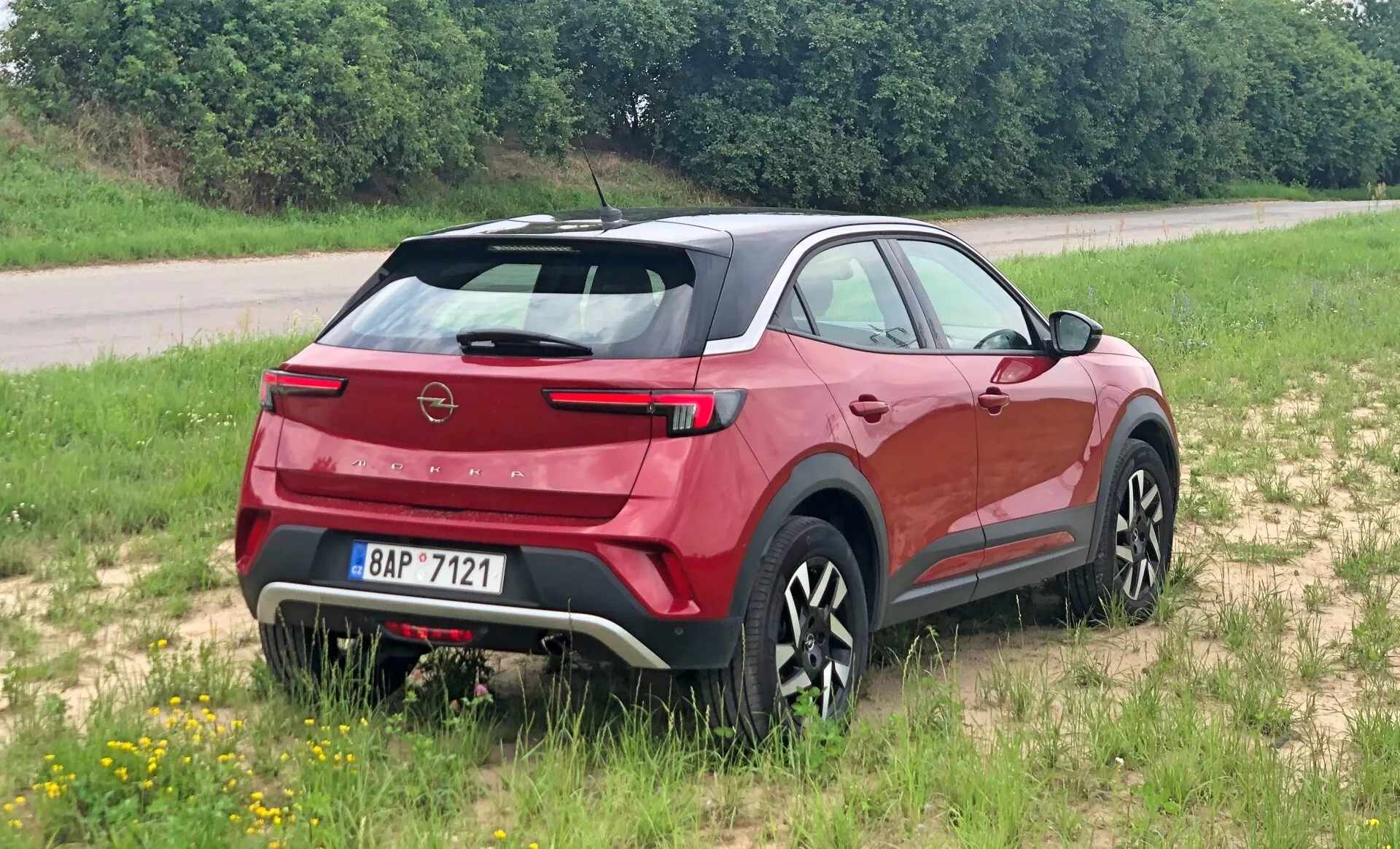 Modern Opel Mokka 2021 crossover SUV hátulról háromnegyedes nézetben, kiemelve a hátsó spoilert, a meredeken döntött hátsó szélvédőt, a jól látható márkajegyet és az elegáns hátsó lámpákat. Füves felületen elhelyezve, jelezve a könnyű terepjáró képességeit. Az oldalsó profil markáns karosszéria vonalakat, fekete burkolatokat és stílusos, többrudazatú kerekeket mutat. Az integrált hátsó ablaktörlő és a parkolási érzékelők, valamint a kontrasztos színű alsó védőlemez az aerodinamikát, a praktikusságot és a robusztusságot sugallják.