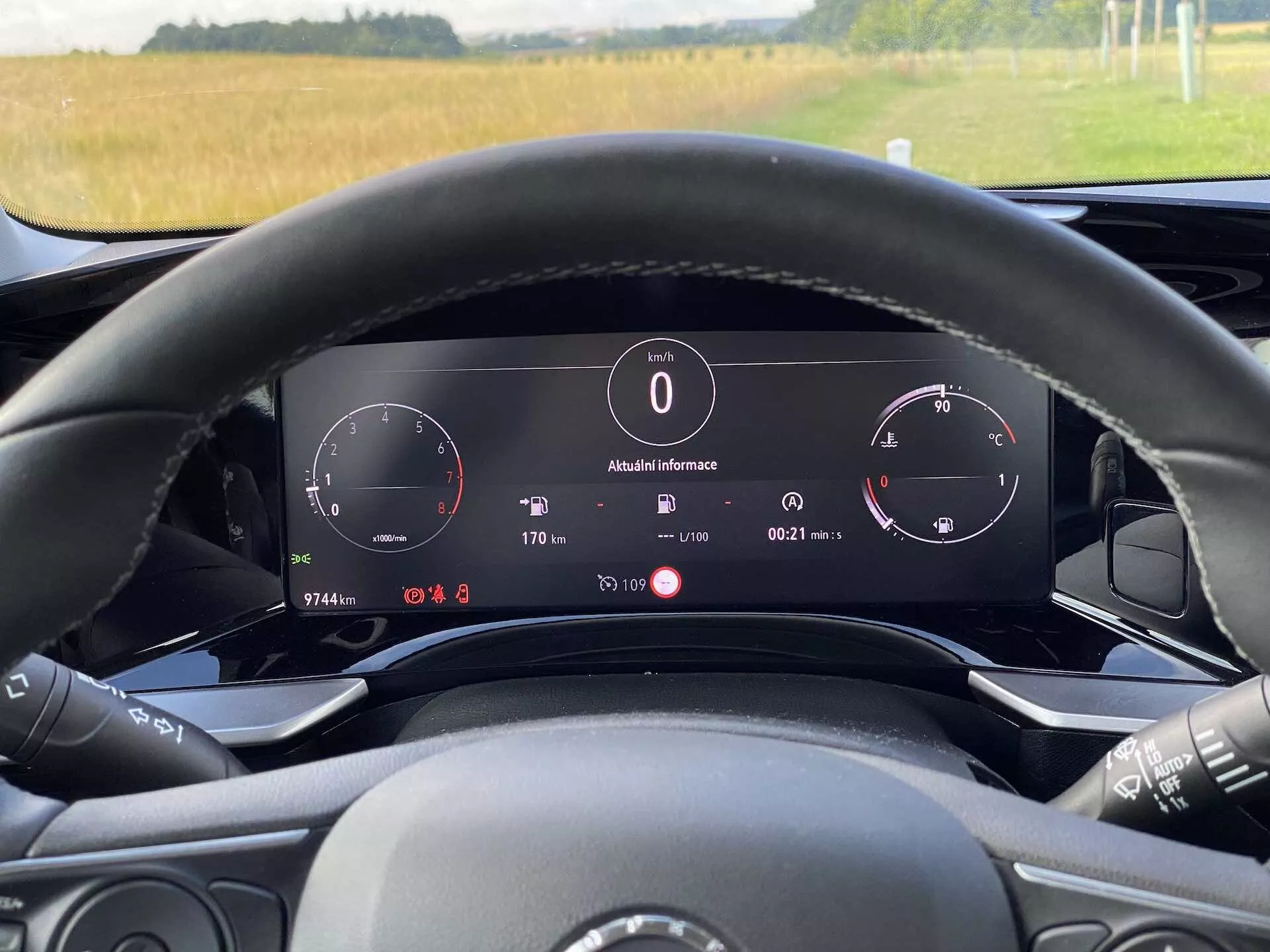 A 2021-es Opel Mokka 1.2 DI Turbo belső nézete a vezető szemszögéből, a kormány mögül. A kép kiemeli a kormány mögötti digitális műszerfalat, amely tartalmazza a sebességmérőt, a fordulatszámmérőt és egy információs panelt, amely megjeleníti a külső hőmérsékletet (8°C), az órát, a sebességváltó pozícióját ('P' parkolóhoz) és az úti számítógép részleteit. A kormánykerék látható média vezérlőkkel rendelkezik és részben látható. A természetes megvilágítás nappali időszakra utal, a szélvédőn keresztül egy vidéki táj látható. Az autó elektronikus kijelzős műszerfalat és fejlett technikai funkciókat mutat be.