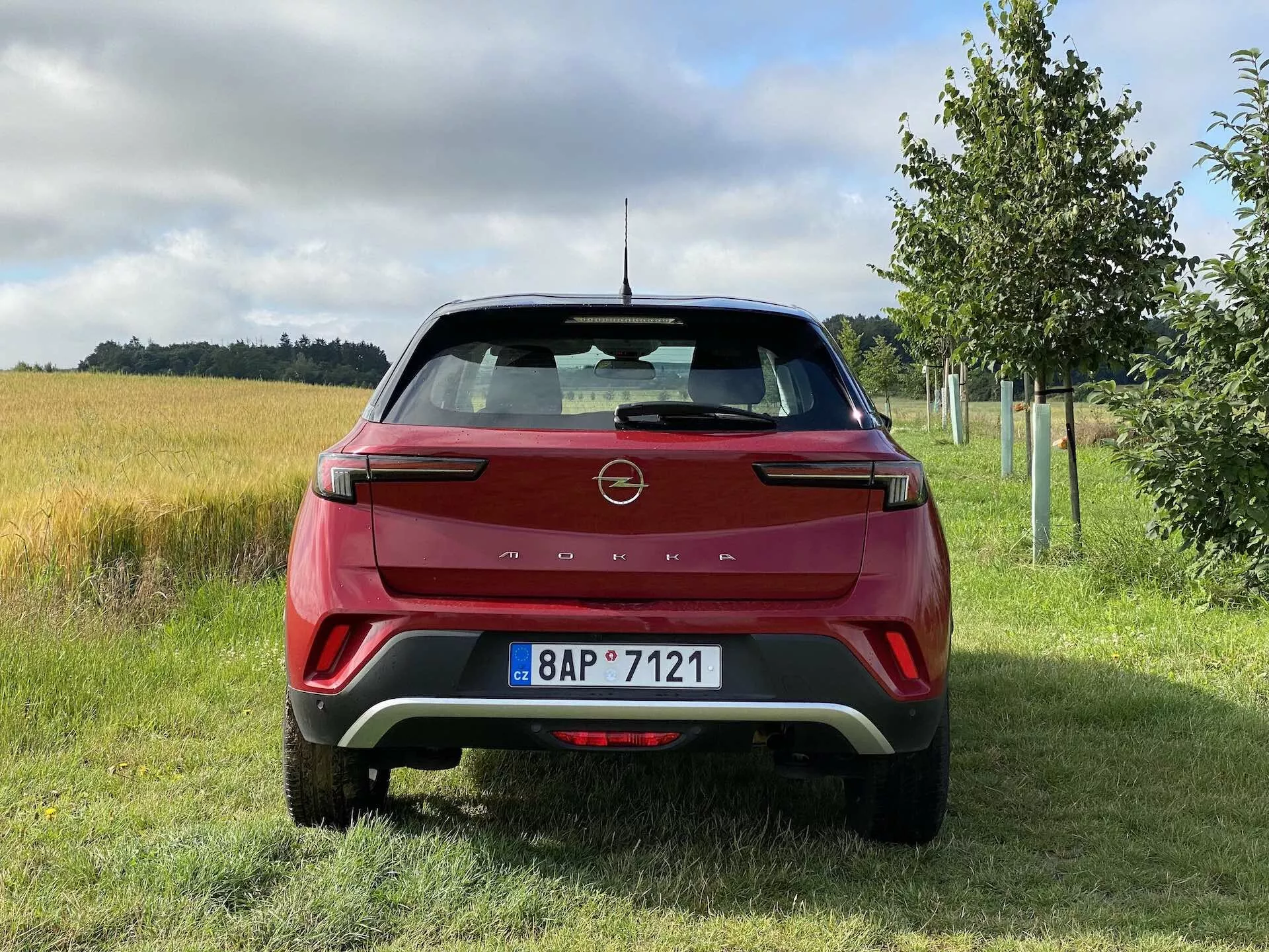 Hátulnézet egy piros 2021-es Opel Mokka 1.2 DI Turbo-ról, modern dizájnnal éles vonalakkal és a gyártó emblémájával a csomagtartón, jellegzetes LED hátsó fényszórókkal, kontrasztos betétekkel ellátott hátsó lökhárítóval, parkolószenzorokkal, integrált fényvisszaverőkkel, hátsó ablaktörlővel, cápauszony antennával, hátsó légterelővel és rendszámtáblával füves, mezőgazdasági területtel és fákkal egy vidéki környezetben.