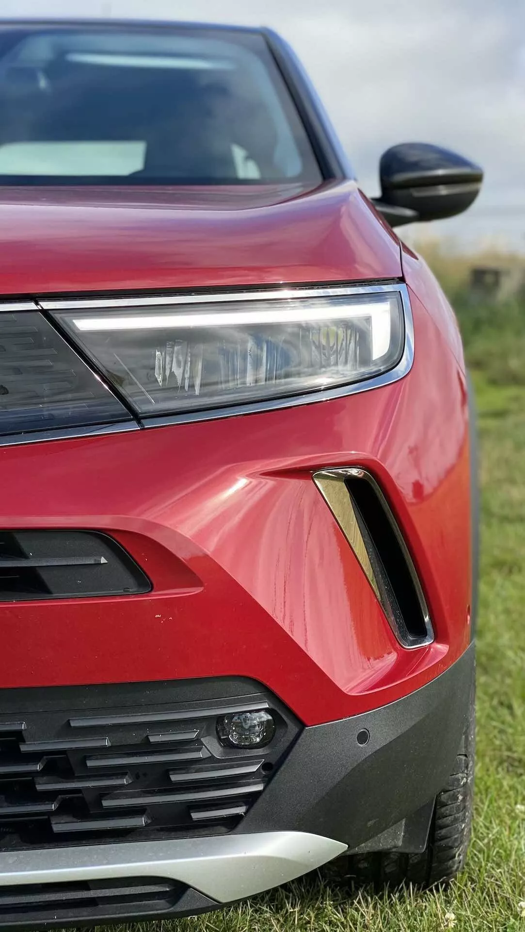 Közelről, szögletes nézet a piros Opel Mokka 2021-es első bal oldaláról. A fotón látható a LED vagy HID világítással ellátott fényszóró, a vízszintes lécekkel rendelkező hűtőrács egy része, a fekete és ezüst kerettel ellátott ködlámpa ház, az élénk piros karosszériafesték erőteljes, szögletes kontúrokkal, az áramvonalas szárnyas tükör és a részben látható motorháztető és sárvédő. Az autó füvön parkol, elmosódott háttérrel.