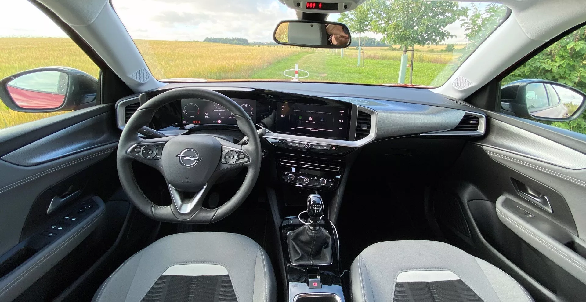 Belső nézet a hátsó ülésből egy Opel Mokka 2021 1.2 DI Turbo-ban. Háromküllős multifunkciós kormánykerékkel, digitális sebességmérő és fordulatszámmérő kijelzővel a kormánykerék mögött, függőleges orientációjú infotainment érintőképernyővel, klímavezérlő kapcsolókkal, manuális sebességváltóval és pohártartókkal, kétszínű sportos első ülésekkel, hozzáillő ajtópanelekkel és kartámasszal, látható visszapillantó tükörrel, részben leengedett ablakokkal és további vezérlőkarokkal a szélvédőtörlőkhöz és az irányjelzőkhöz.