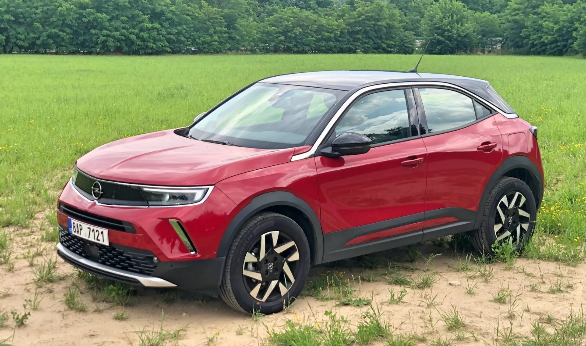 Kép egy 2021-es piros Opel Mokka SUV-ról, amely füves felületen parkol a háttérben mezővel. Ez a háromnegyedes elülső nézet kiemeli a hűtőrácsot, ami potenciálisan fejlett hajtáslánc-technológiára utal, elegáns LED-es nappali menetfényt, ködlámpákat és egyedülálló kéttónusú alufelniket. Láthatók a karosszéria színére festett visszapillantó tükrök és kilincsek, a dinamikus vállvonal, a védő fekete alsó díszléc és a krómozott ablakkeretek is, bemutatva a jármű modern és luxus dizájn jellemzőit.