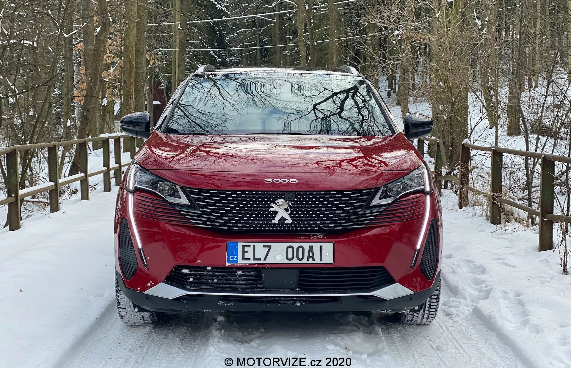 Vörös Peugeot 3008 2021 elölnézete havas úton parkolva, jellegzetes hűtőrács mintával, központi Peugeot emblémával, éles első lámpákkal és agyarszerű nappali menetfénnyel, kontúros motorháztetővel, első lökhárítóba integrált parkolószenzorokkal és havas erdei környezettel.