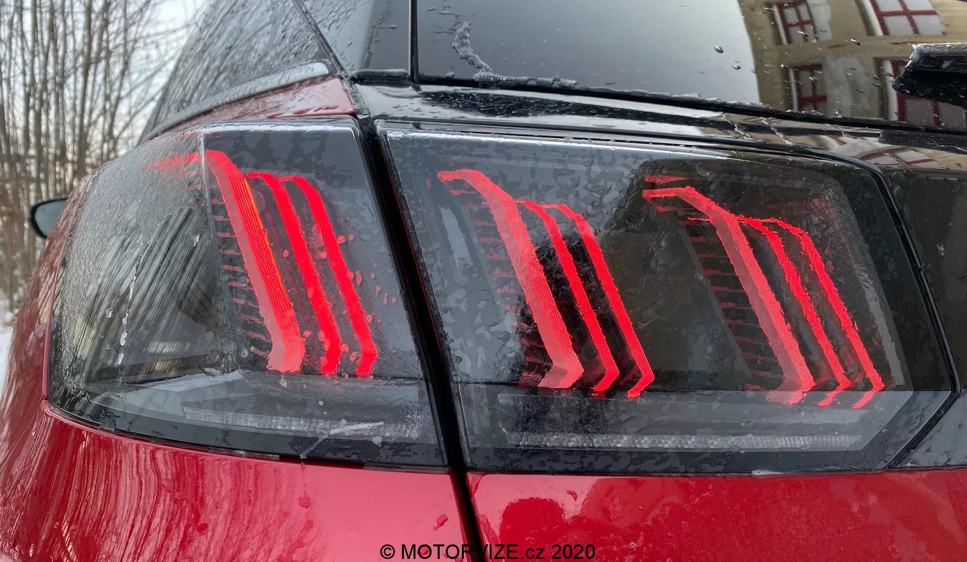 Közelkép egy 2021-es Peugeot 3008 hátsó részéről, kiemelve a modern, függőlegesen világító LED hátsó lámpa dizájnját a fényes vörös karosszérián vízcseppekkel, ami eső közbeni vagy utáni kültéri környezetet sugall; a hátsó lámpa energiahatékony technológiával rendelkezik, és féklámpa és helyzetjelző lámpaként is funkcionál.