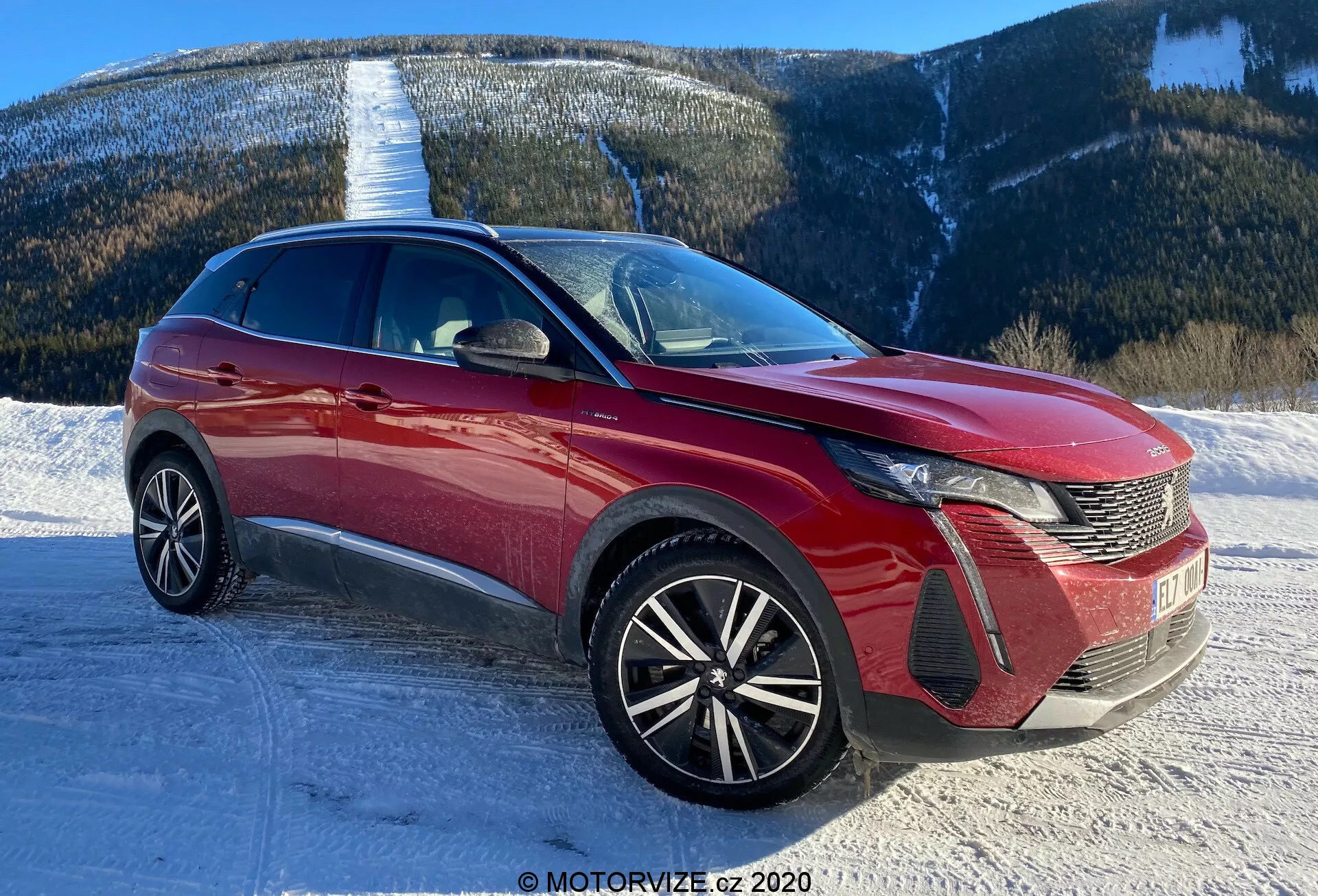 Kép egy modern autóról, valószínűleg egy 2021-es Peugeot 3008-ról, elülső háromnegyedes perspektívából fényképezve. A szög jól mutatja az autó jellegzetes LED-fényszóróit, a fésűmintás hűtőrácsot és az első emblémát. Az első lökhárítónak légbeömlős részei és ködlámpatokjai vannak. A motorháztető formázott vonalakból áll, amelyek hozzájárulnak az aerodinamikához, míg a széles szélvédő jó előre látogatóságot biztosít. A sokküllős könnyűfém keréktárcsák sportosabb megjelenést és magasabb felszereltségi szintet sugallnak. A karcsú tetővonal és a dinamikus oldalsó vonalak crossoverre vagy SUV-ra utalnak. Láthatóak az integrált irányjelzős oldalsó tükrök és a karosszéria alsó oldalán lévő díszítés. A helyszín havas úton, hegyi háttérrel azt sugallja, hogy a jármű különböző terepeken is megállja a helyét.