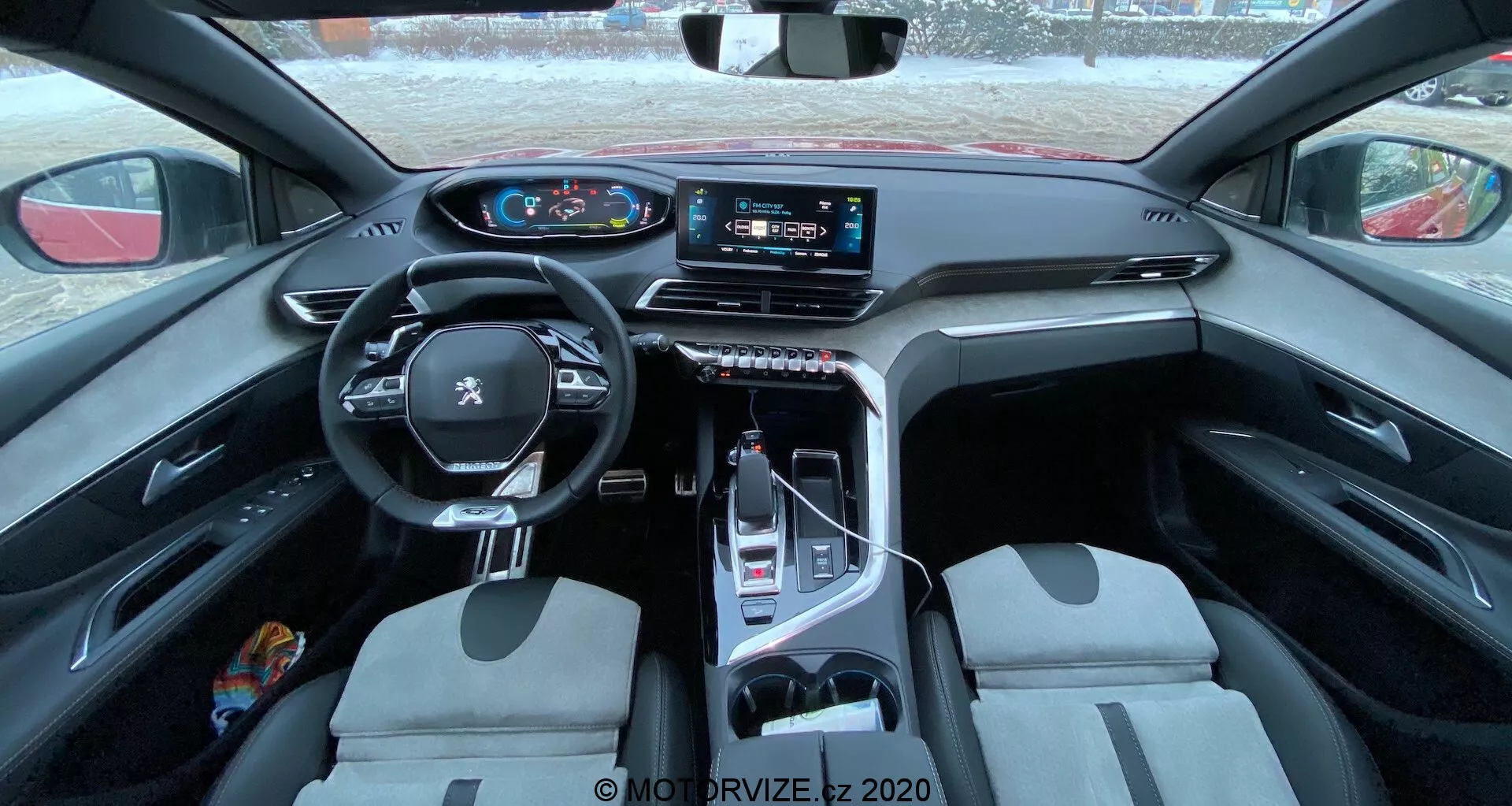 A 2021-es Peugeot 3008 belső nézete, amelyen egy többfunkciós, bőrborítású kormánykerék látható beépített vezérlőelemekkel, digitális műszercsoport, nagy érintőképernyős információs és szórakoztatórendszer a középkonzolon, elektronikus sebességváltó választó, kétszínű kialakítású első ülések és praktikus és luxus anyagok keveréke. A kép a hátsó ülésből készült, tágas bepillantást nyújtva az utastérbe, kiemelve a dizájnt és a technológiai jellemzőket.