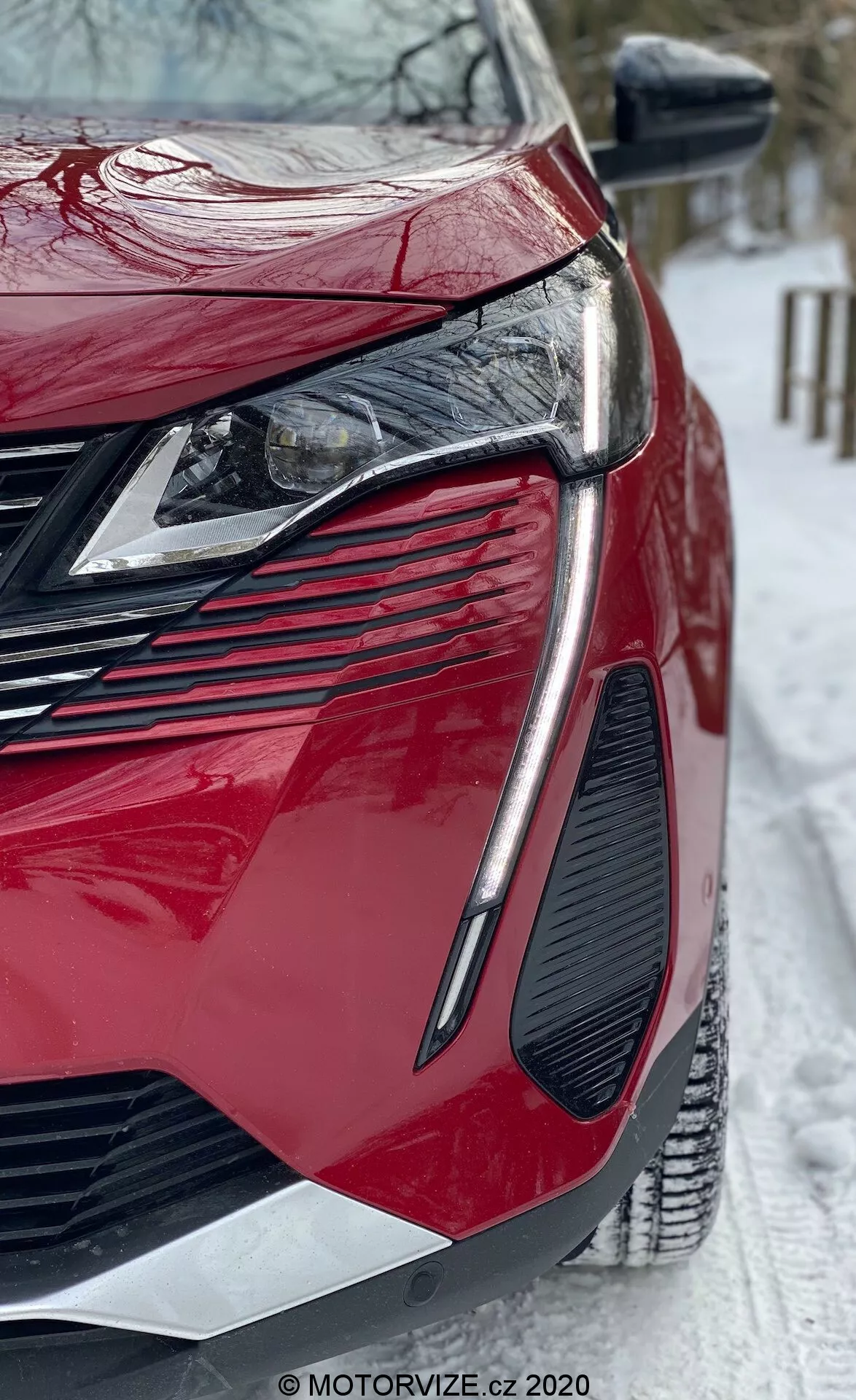 Modern, piros Peugeot 3008 2021 közelkép alacsony szögből. Az elülső-bal negyedre fókuszálva, bemutatva elegáns, szögletes LED fényszórókat, nagy, agresszív hűtőrácsot, jellegzetes szellőzőnyílás-kialakítást, fényes festékbevonatot és élesen kontúrozott karosszériapaneleket. Az oldalsó tükörbe irányjelzők vannak integrálva, és a jelenet havas talajt tartalmaz, téli körülményeket sugallva. MOTORVIZE.cz felirat látható.